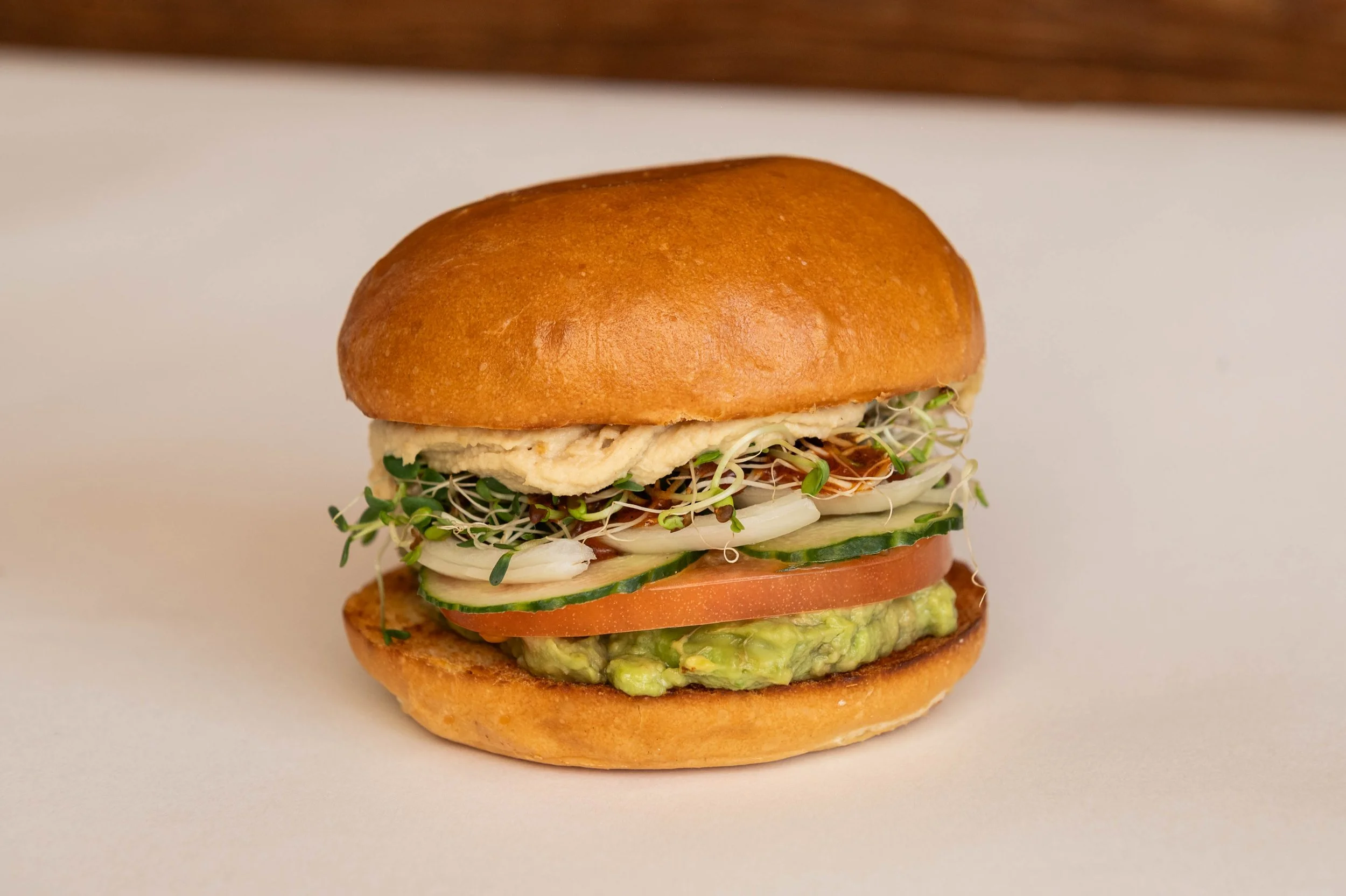 A sandwich with scrambled eggs, cucumber slices, tomato slices, guacamole, sprouts, and onion slices in a bun.