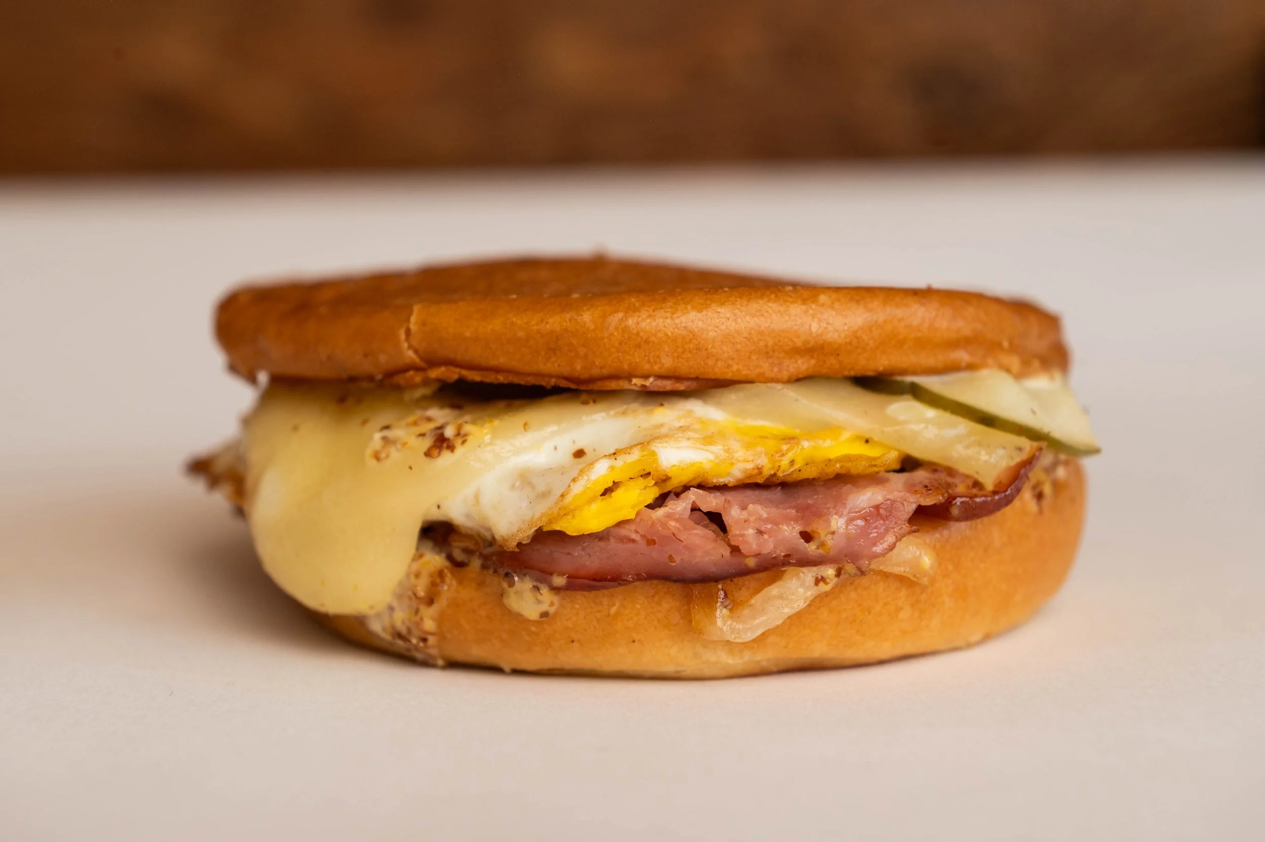 Close-up of a breakfast sandwich featuring melted cheese, fried egg, bacon, and pickles on a toasted bun.