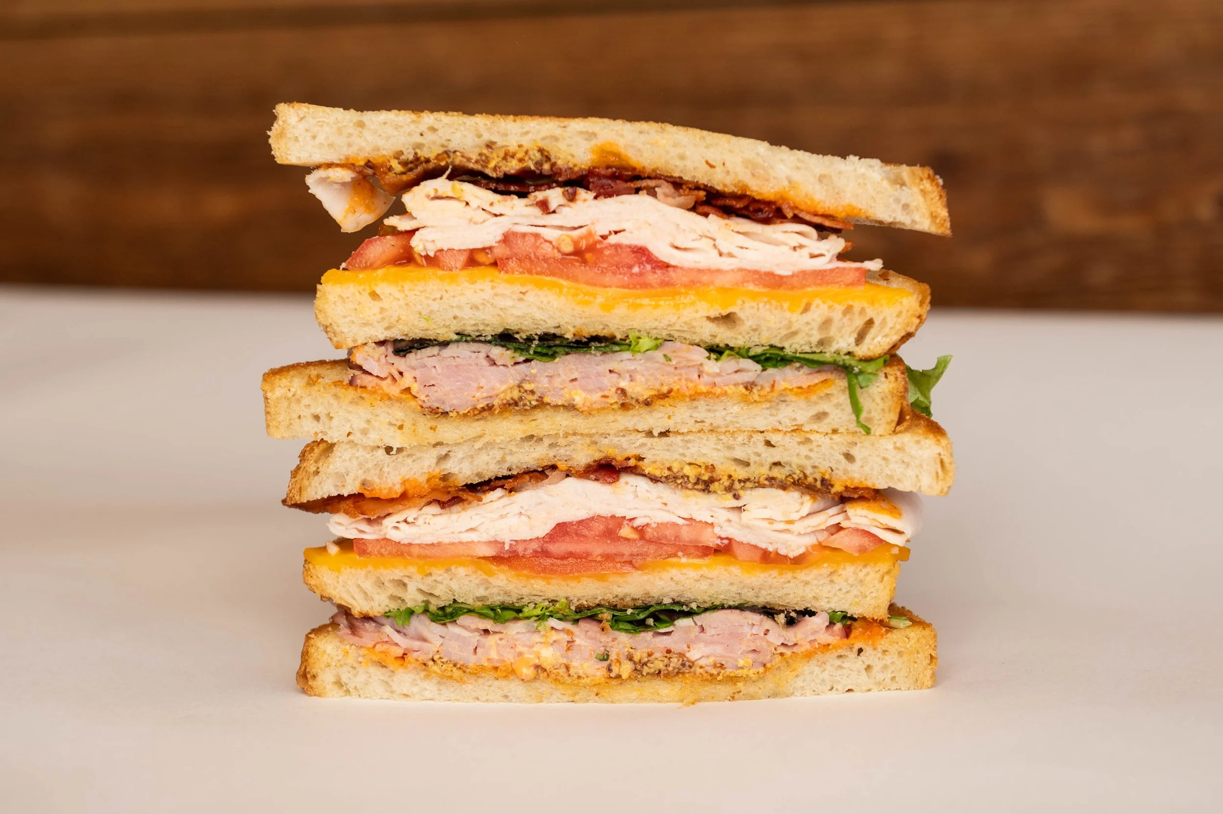 A stack of four turkey sandwiches with lettuce, tomato, cheese, and bread, on a white surface with a wooden background.