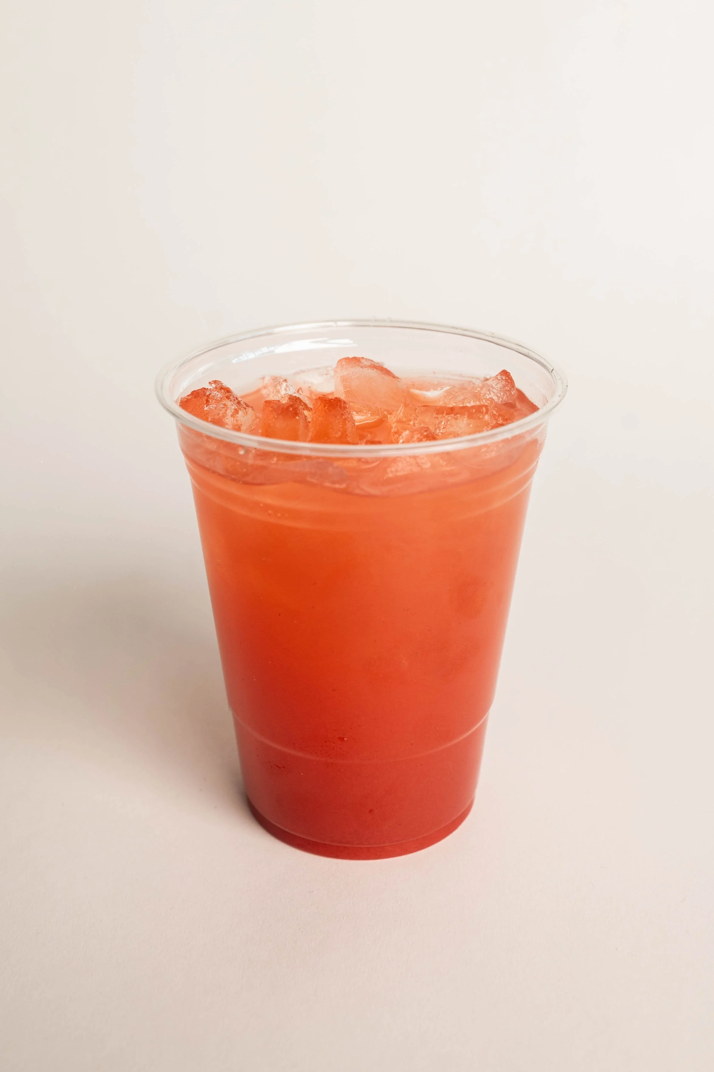 A clear plastic cup filled with pinkish-red beverage and ice cubes, set against a plain white background.
