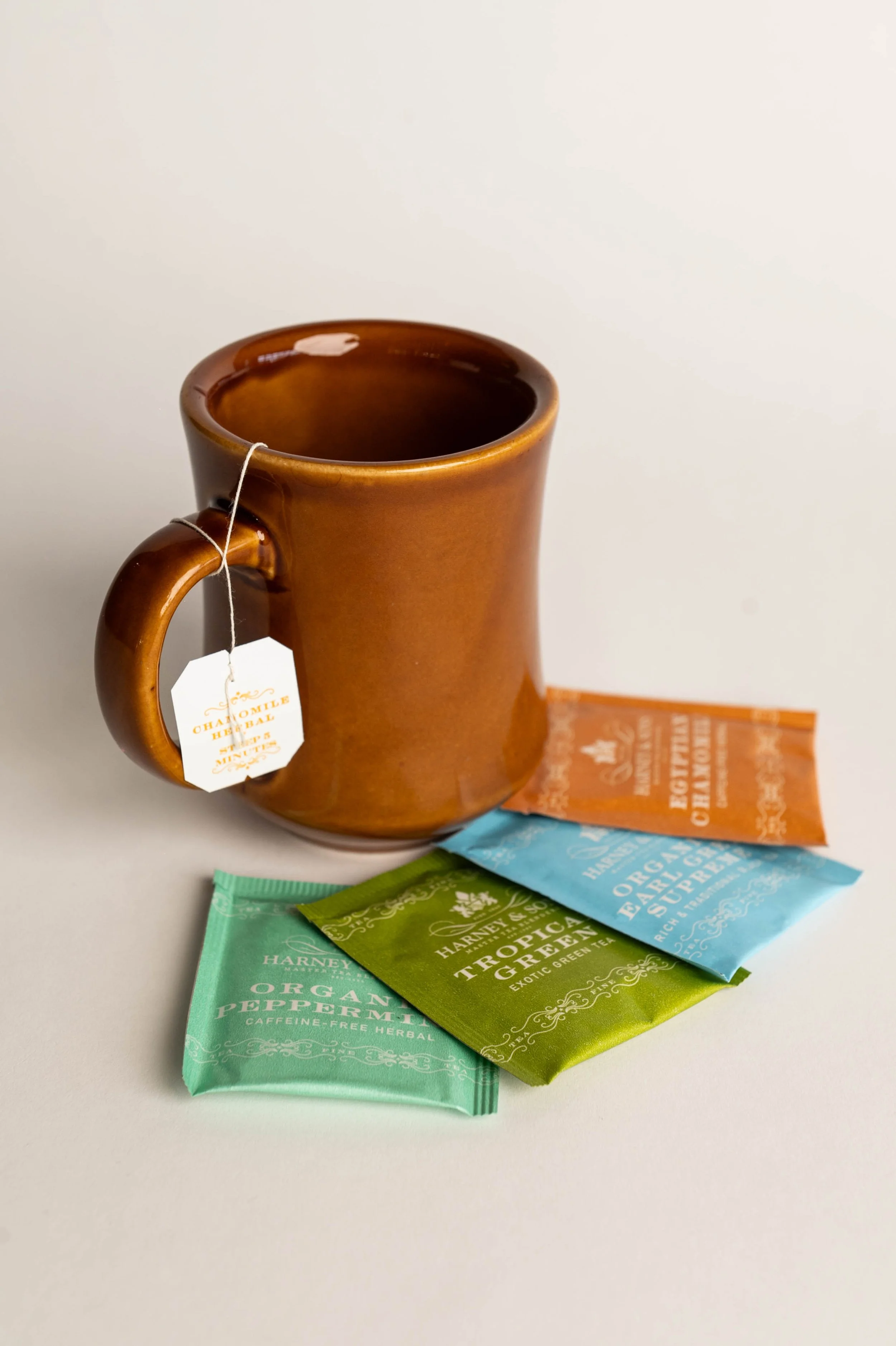 A brown ceramic mug with a tea bag hanging from the handle and three colorful tea sachets on a white surface.