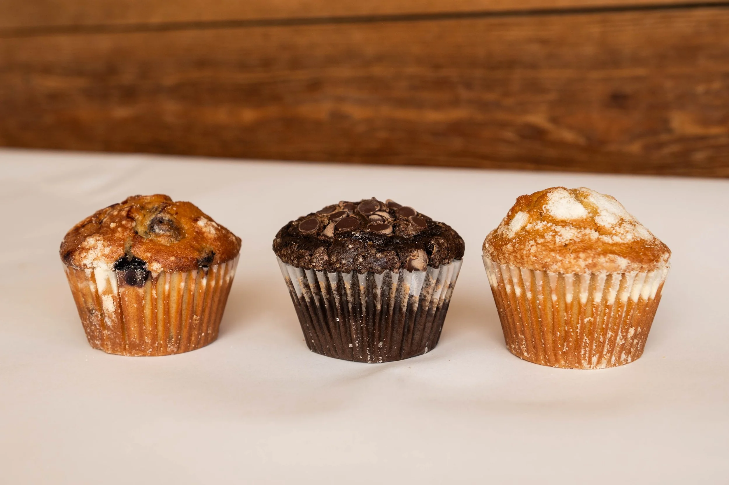 Three muffins arranged in a row on a white surface, with a wooden background. The muffin on the left has chocolate chips and a dusting of powdered sugar. The middle muffin is chocolate, topped with chocolate chips. The muffin on the right is light-co