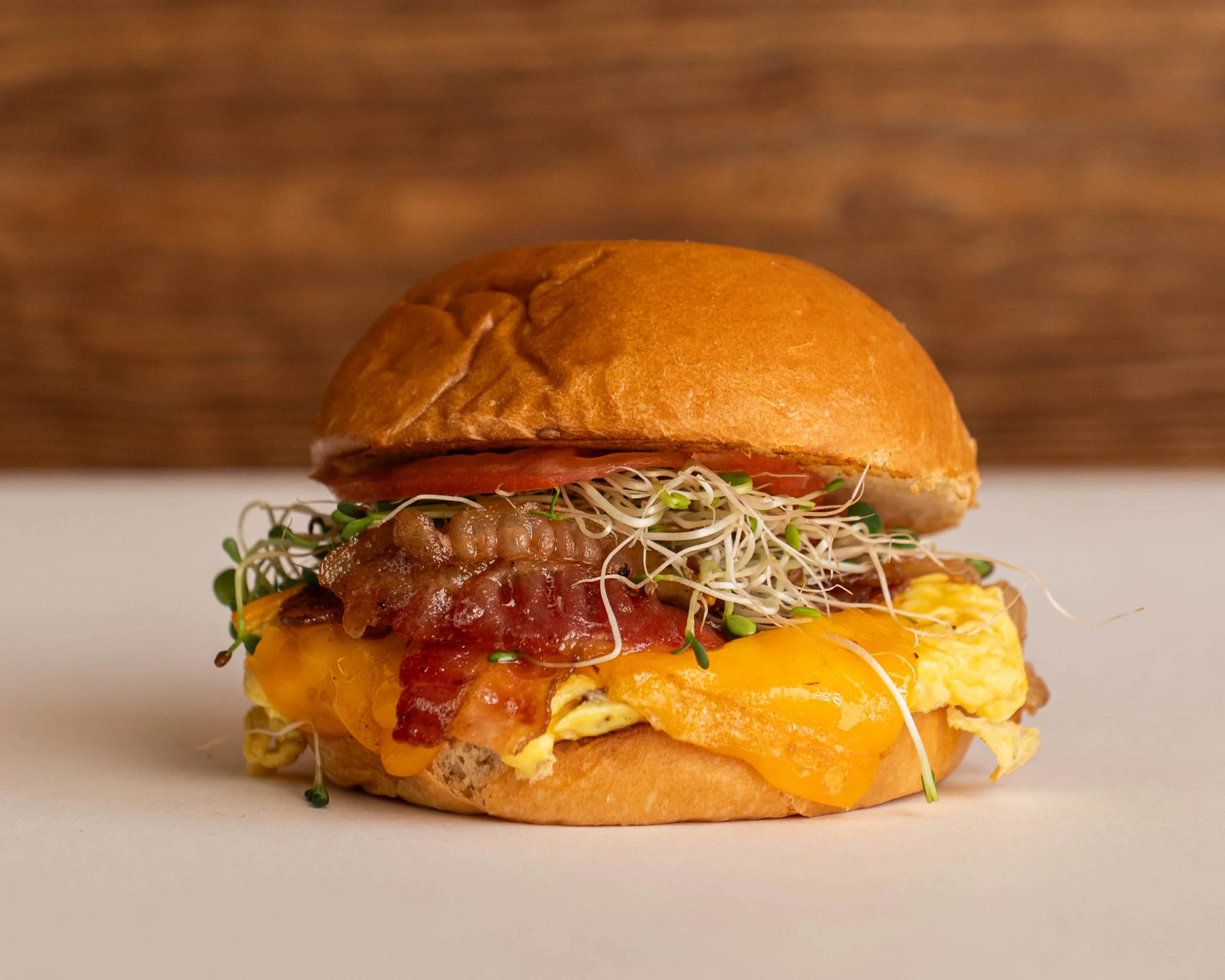 Close-up of a breakfast sandwich with scrambled eggs, cheese, bacon, sprouts, tomato, and a toasted bun.