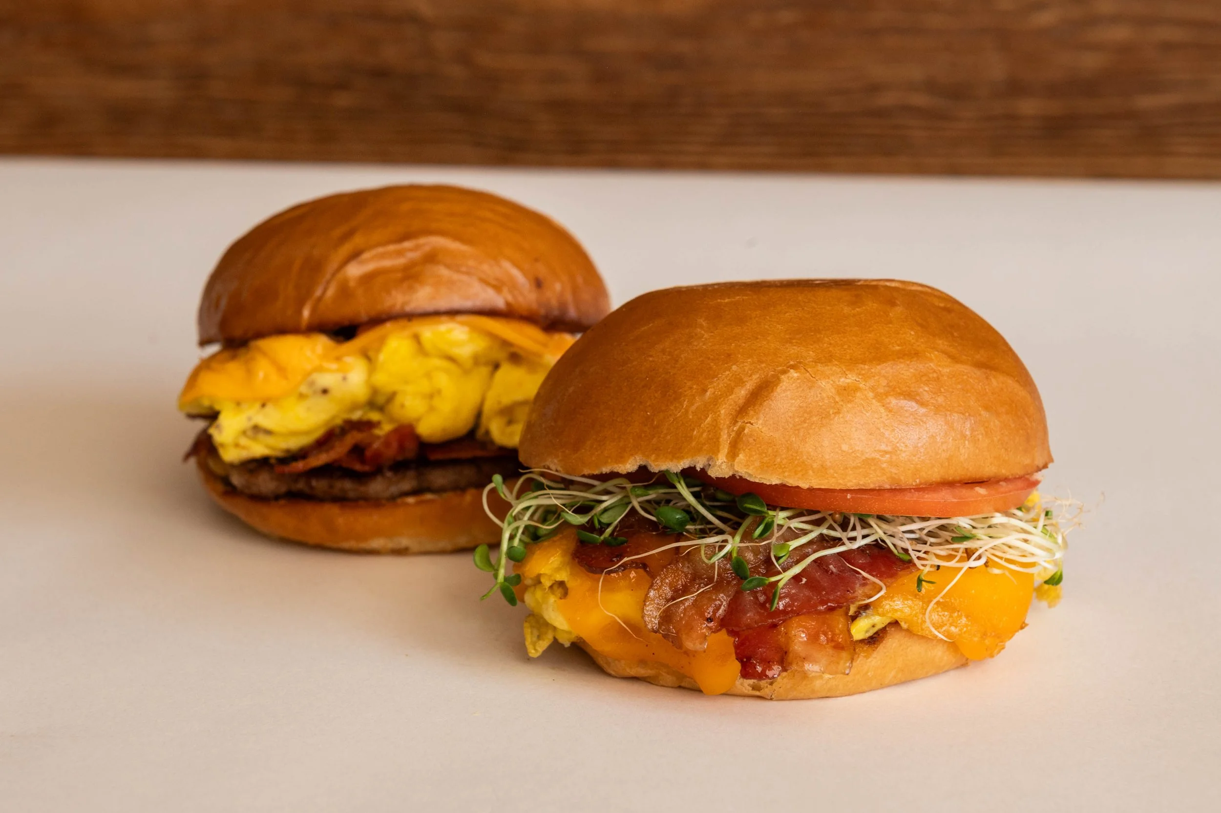 Two breakfast sandwiches on a white surface with a brown background. The sandwich in the foreground has a bun, scrambled eggs, cheese, bacon, tomato, sprouts, and additional ingredients. The sandwich in the background has a bun, scrambled eggs, cheese, and possibly bacon.
