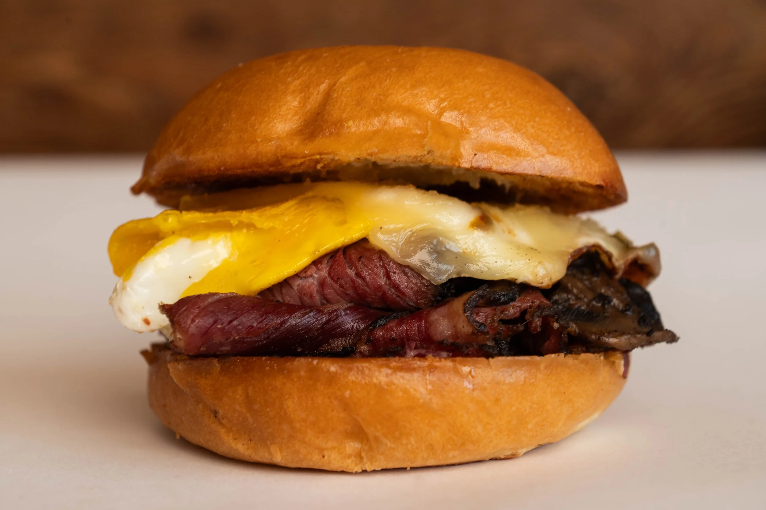 Close-up of a breakfast sandwich with a toasted bun, fried egg, slices of roast beef, and a beef patty.