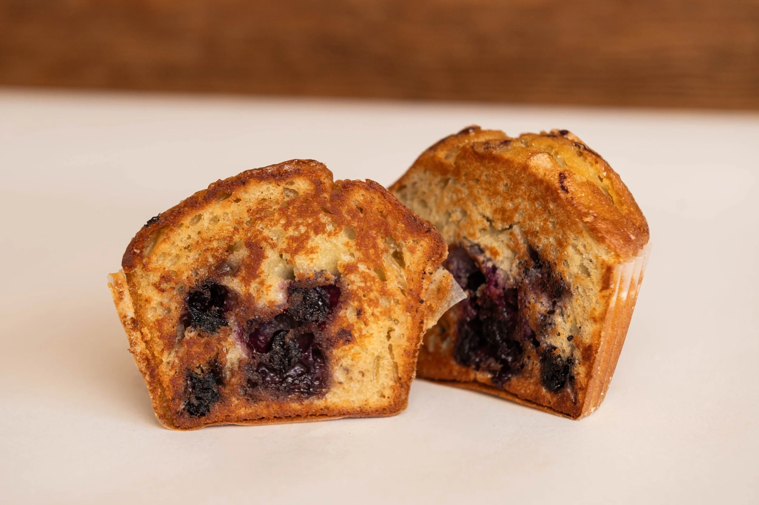 Close-up of two blueberry muffins broken in half, showing blueberries inside, on a white surface with a wooden background.