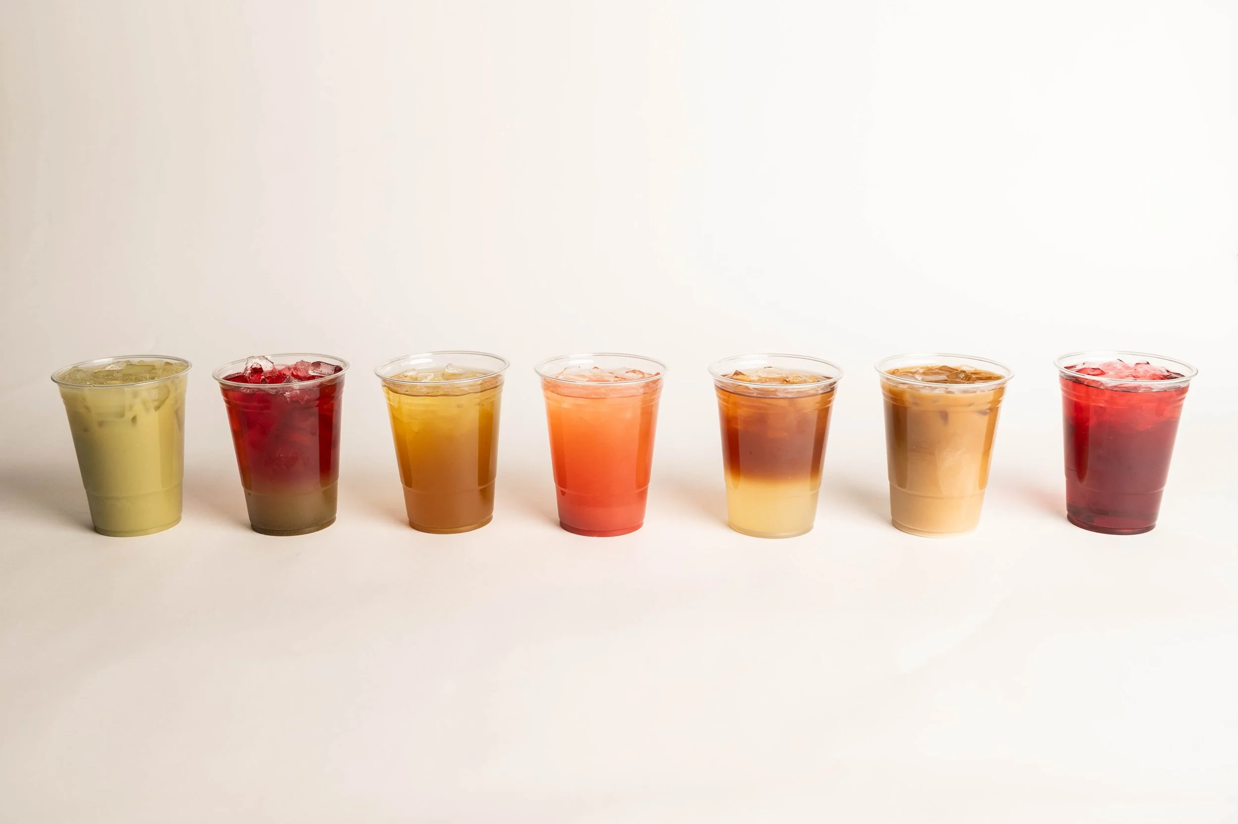 Seven cups of iced beverages with varying colors from yellow to red, lined up on a white surface against a plain white background.