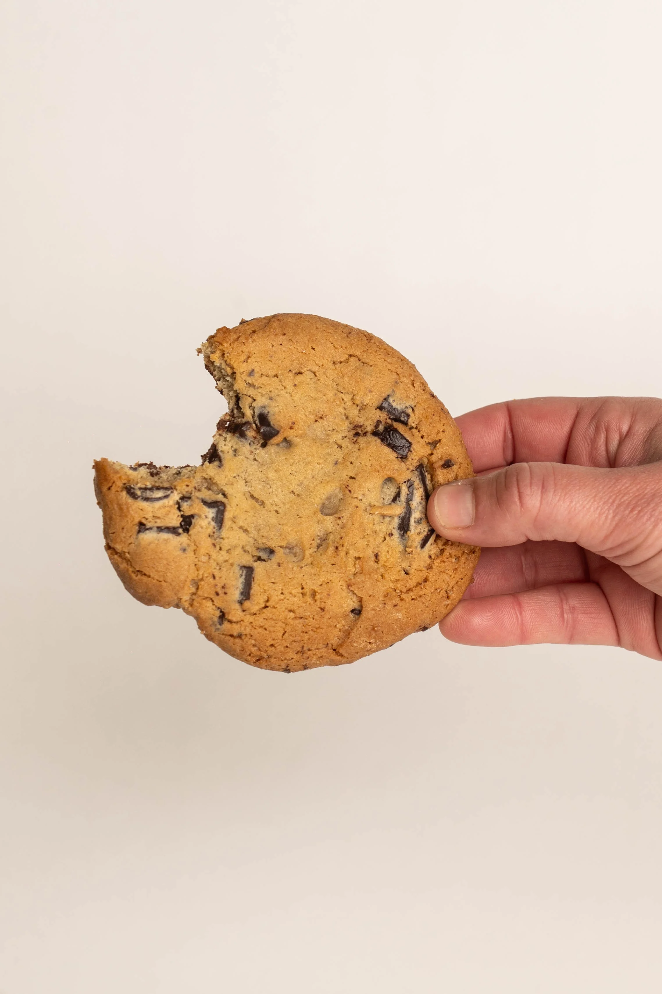 Hand holding a partially eaten chocolate chip cookie against a plain, light-colored background.