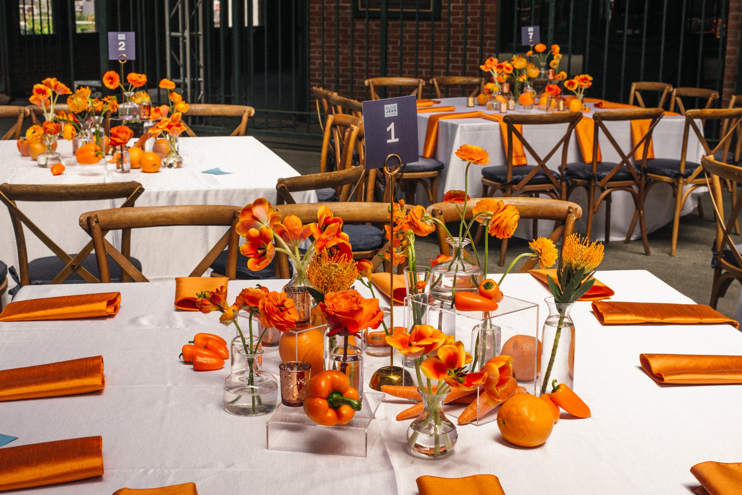 Table decorations featuring orange flowers, fruits, and vegetables arranged in glass vases and containers at a banquet or event.