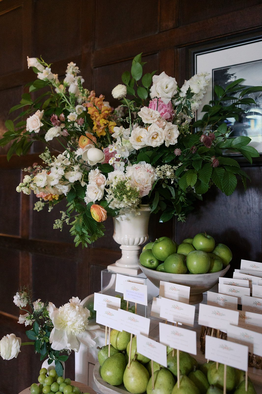 A large floral arrangement with white and pastel-colored flowers in a white vase, with green apples and white name cards beneath, set against a dark wood-paneled wall.