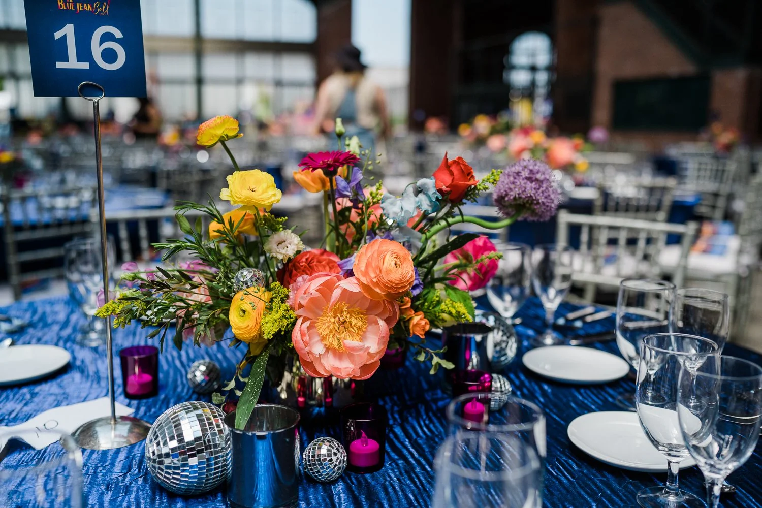 Decorated banquet table with colorful floral centerpiece, glassware, plates, and small pink and disco ball decorations, set in a venue with a blue ruched tablecloth.