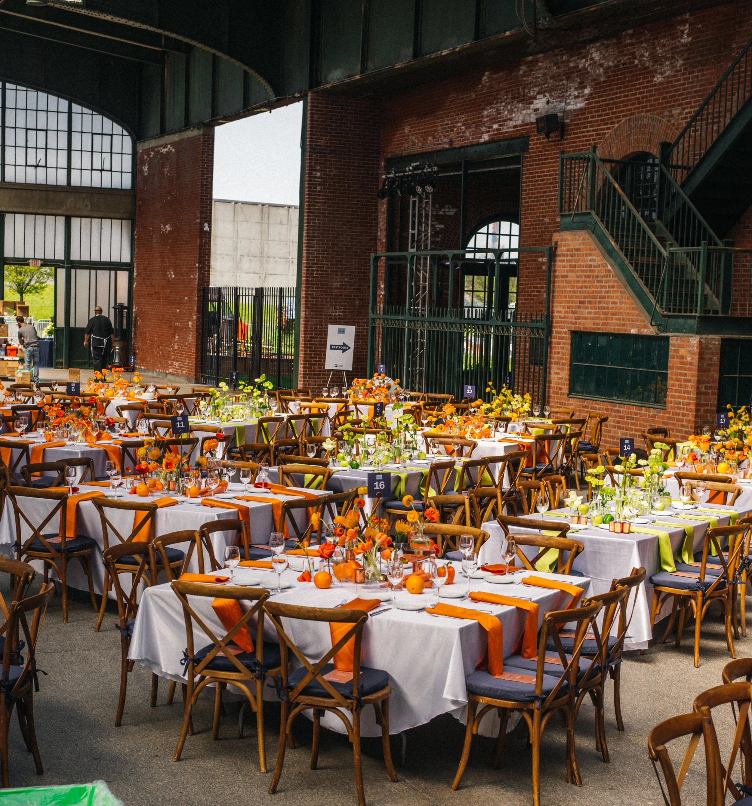 Indoor event space set for a celebration with decorated tables, orange and green centerpieces, and floral arrangements in a rustic, industrial-style venue with exposed brick walls and large windows.