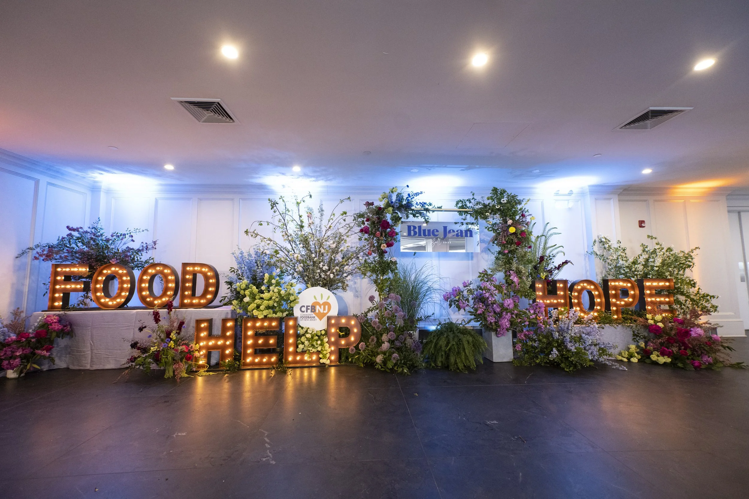 Decorative event display with illuminated signs reading 'FOOD HELP' and 'HOPE', surrounded by floral arrangements and a sign for Blue Jean.