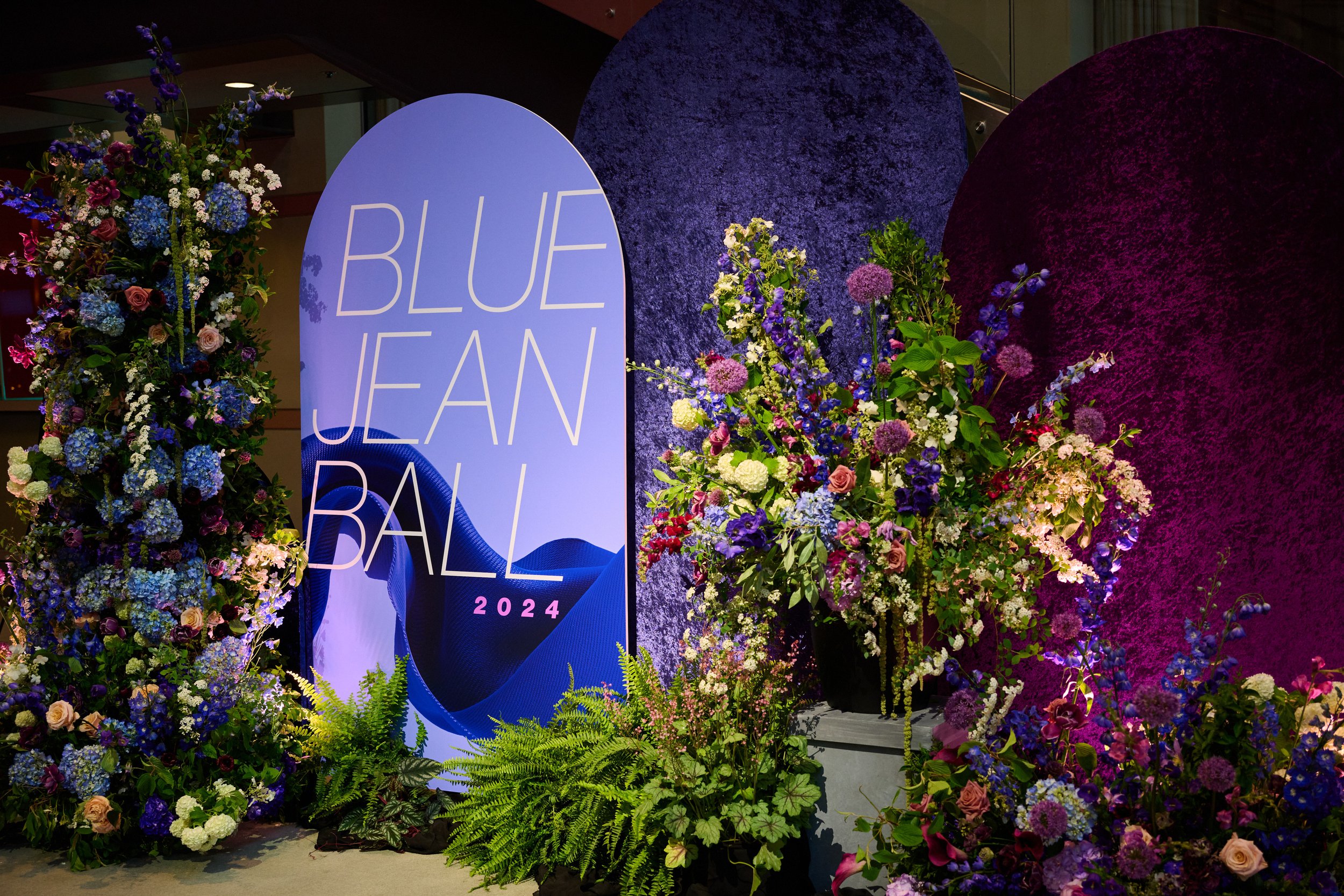 Floral display featuring purple, pink, white, and blue flowers with a large blue sign reading "Blue Jean Ball 2024" at an event.