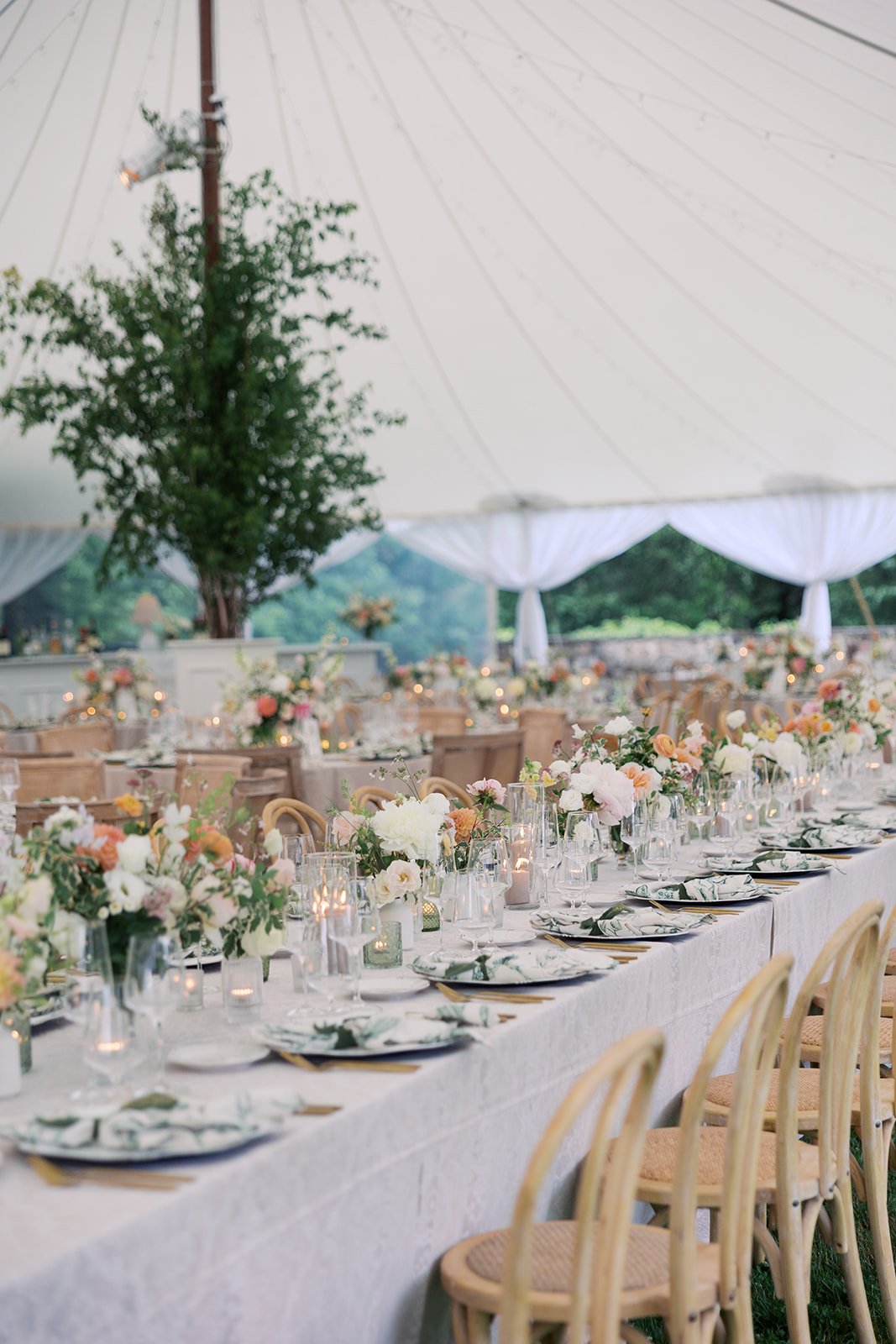 Elegant outdoor wedding reception tent with long table decorated with white linen tablecloth, floral centerpieces in soft pastel colors, candles, and set with plates, napkins, and glassware, surrounded by wooden chairs.