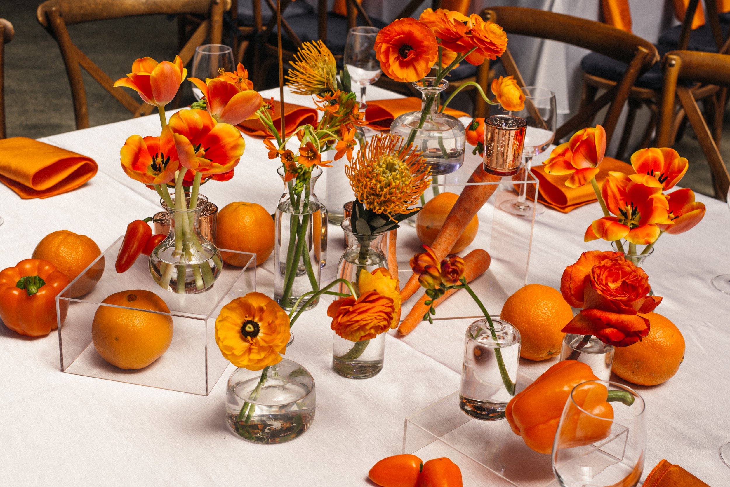 A table decorated with orange flowers in vases, oranges, and orange peppers, with orange napkins and glasses set for a meal.