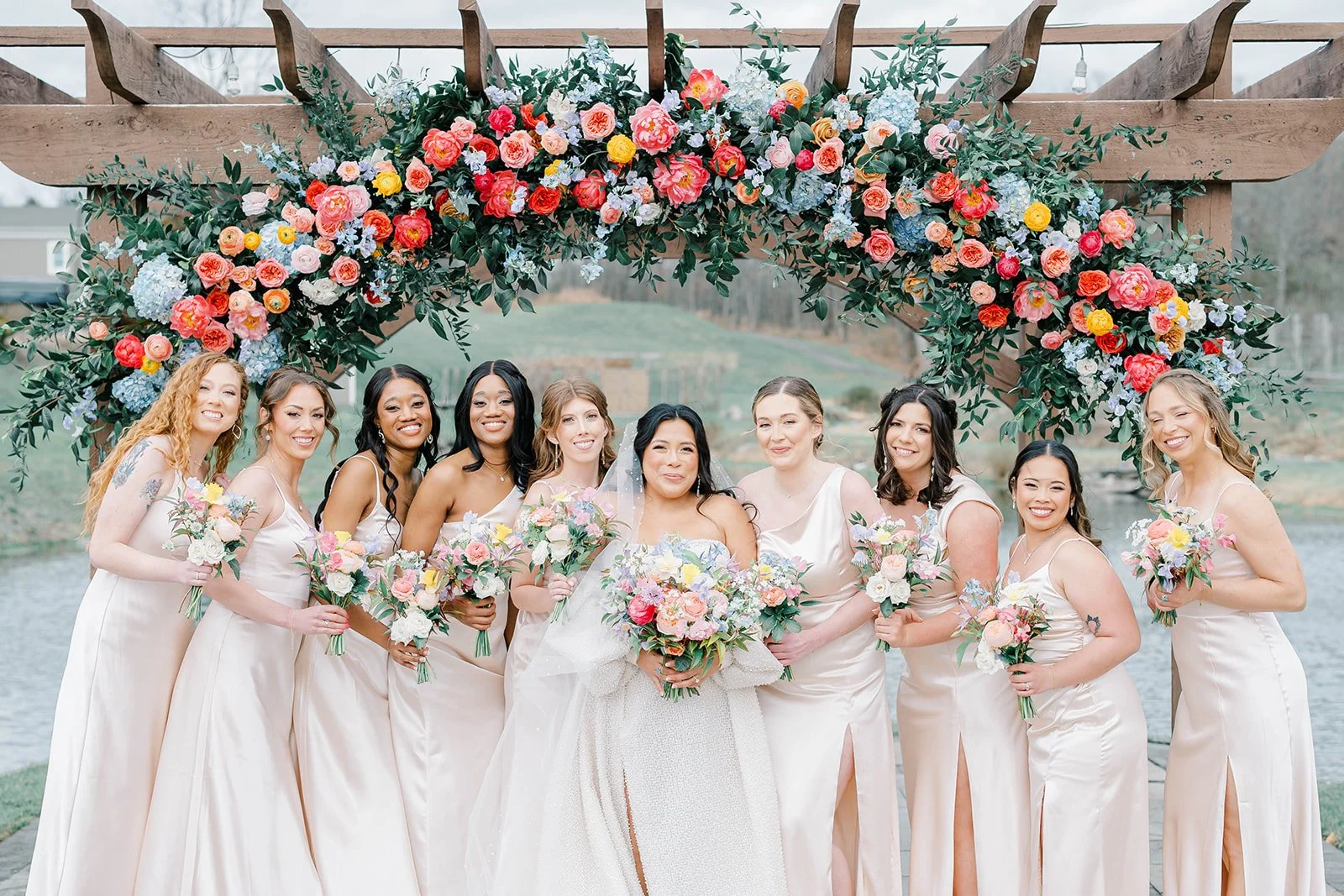 A bride and nine bridesmaids standing under a floral arch by a river, all dressed in light-colored gowns holding bouquets, smiling for a photo.
