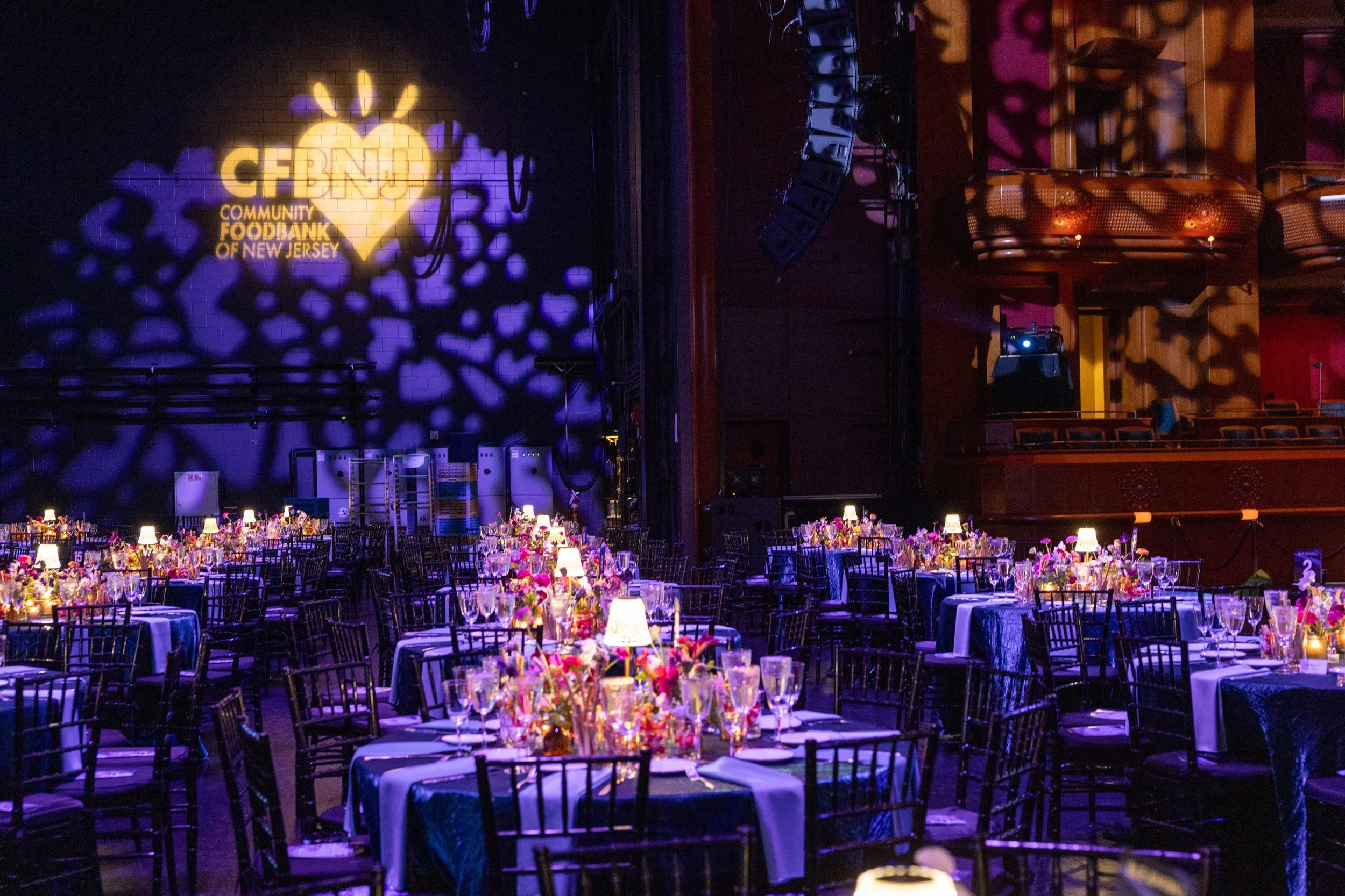 Dimly lit event hall with decorated tables, floral centerpieces, and small lamps, with a large illuminated wall projection reading 'CFBNJ Community FoodBank of New Jersey'.