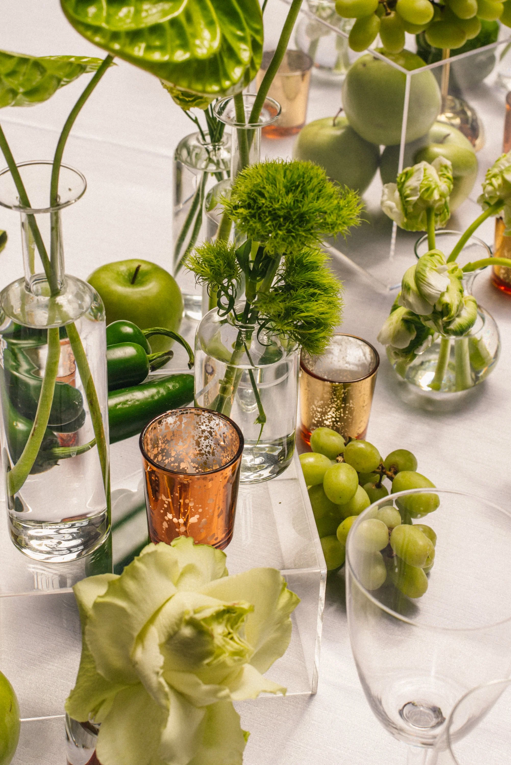 Green apples, grapes, white flowers in glass vases, and a few decorative glasses on a white tablecloth.