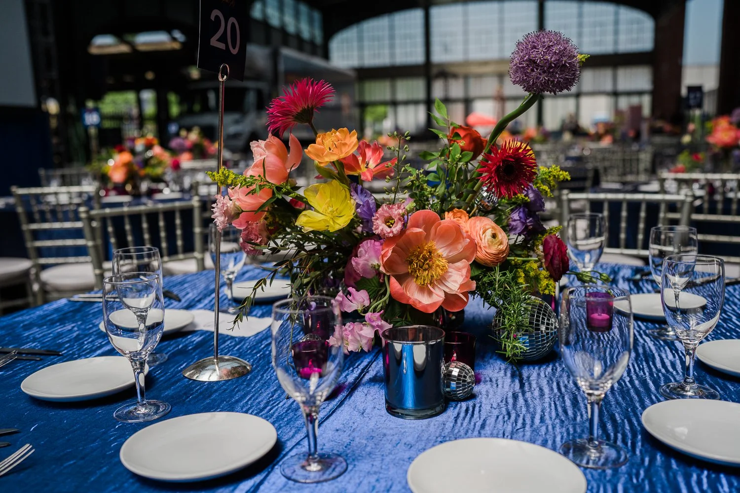 A banquet table decorated with a large colorful floral centerpiece, surrounded by empty white plates, wine glasses, and purple candles, set in a bright indoor space with high windows.