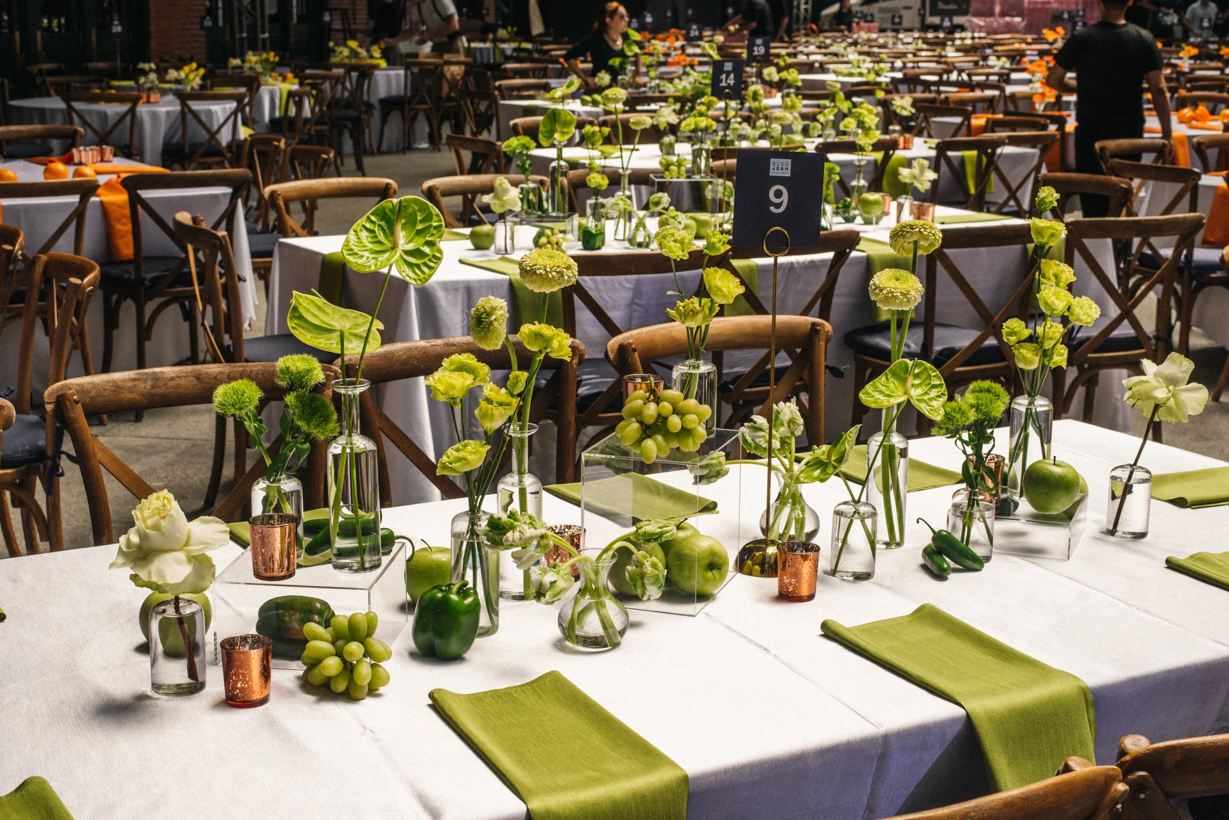 Formal event setup with long tables decorated with white tablecloths, green floral arrangements, and green-themed accents, in a large banquet hall.