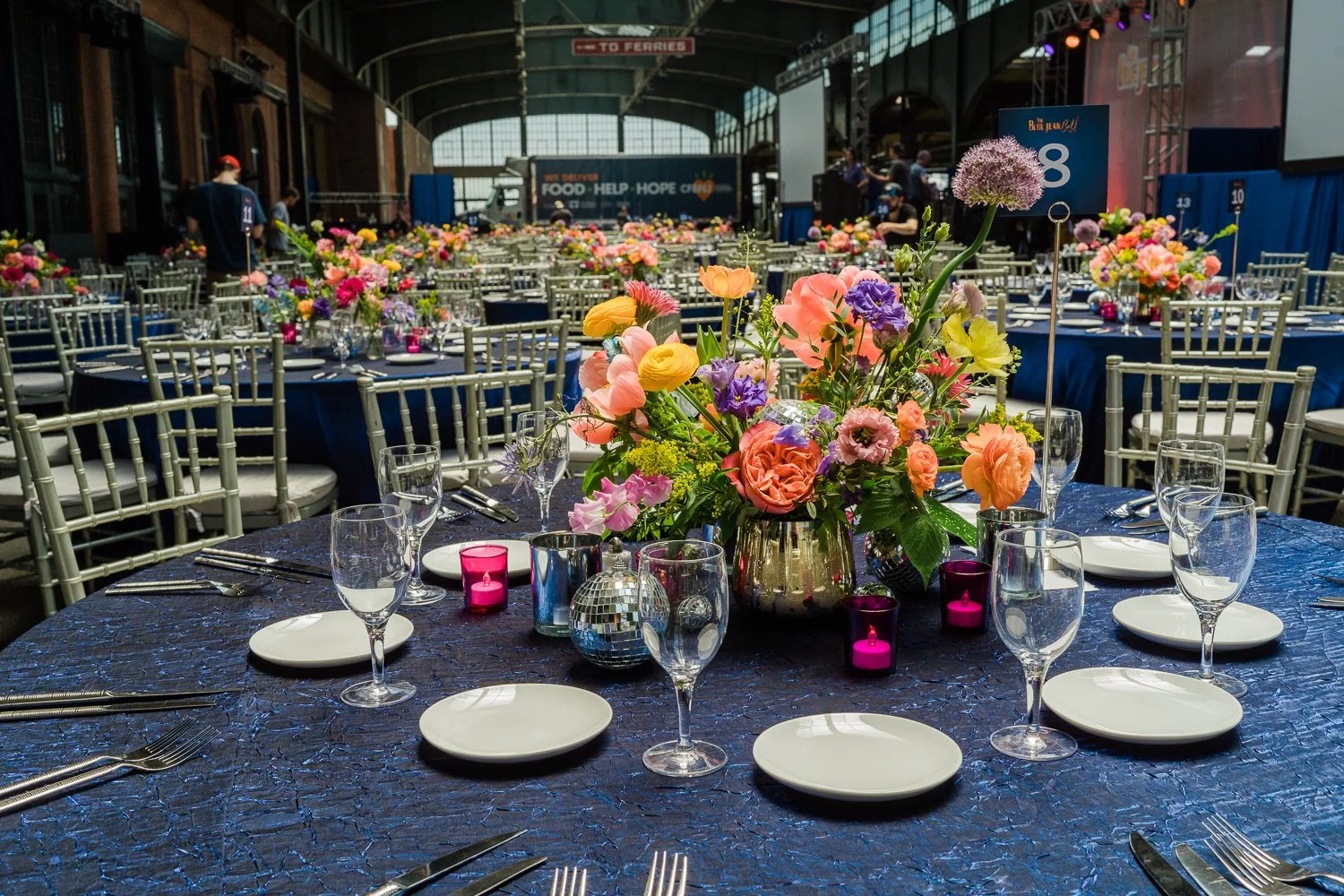 Elegant banquet table decorated with colorful floral centerpieces, wine glasses, and candles, set in a large indoor venue with multiple similar tables and a stage in the background.