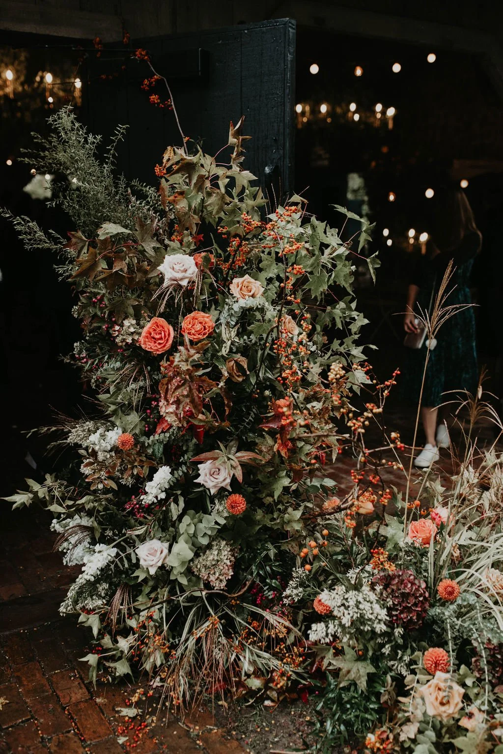 A large floral arrangement with roses, greenery, and berries set against a dark background with string lights and a person in the distance.