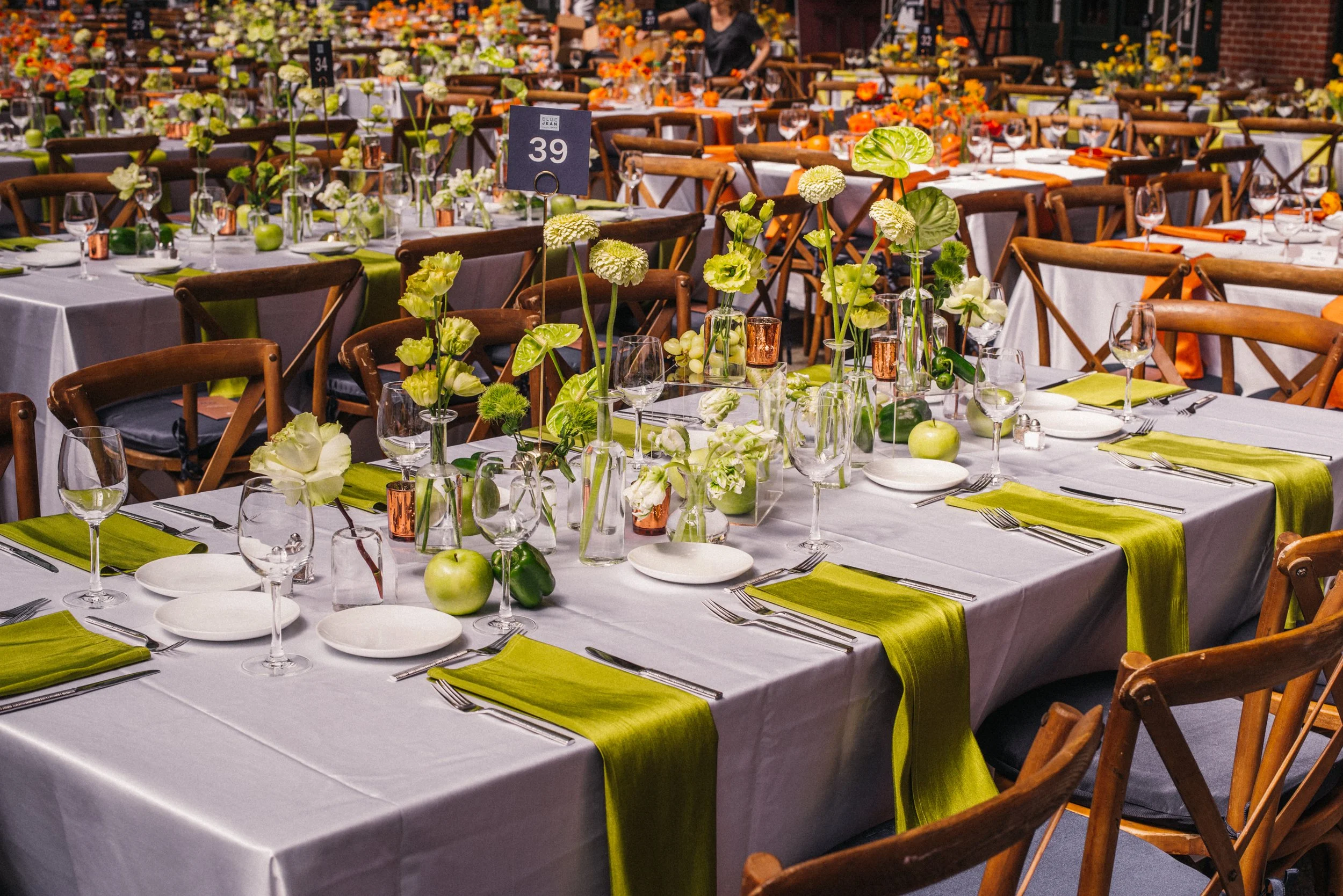 Wedding reception table decorated with white tablecloths, green napkins, and floral centerpieces in glass vases, set for multiple guests with plates, silverware, and glasses, with one table numbered 39.