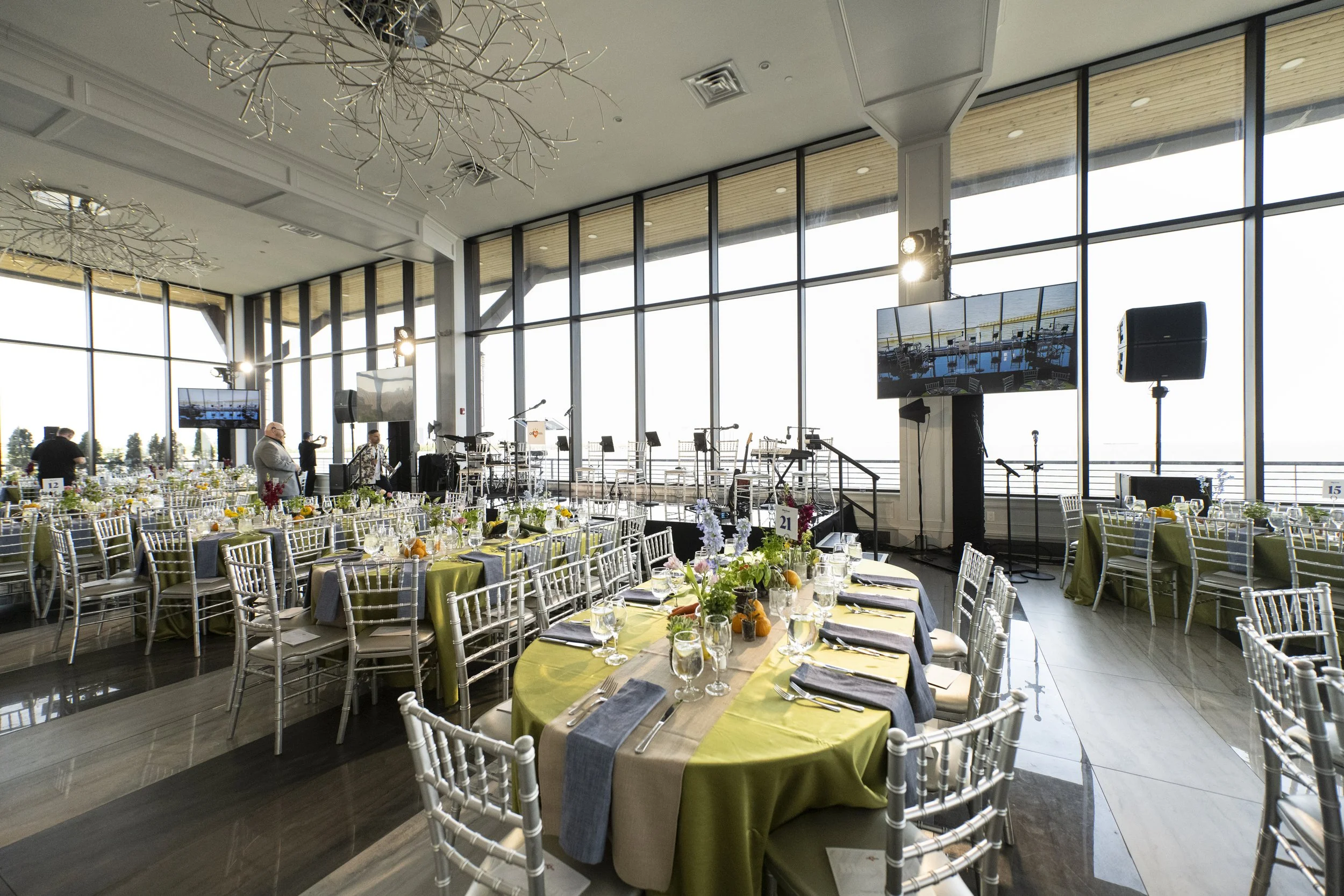Elegant event space with round tables set for a formal dinner, decorated with flowers and orange accents, and an elevated stage area with musical instruments and large screens, surrounded by floor-to-ceiling windows.