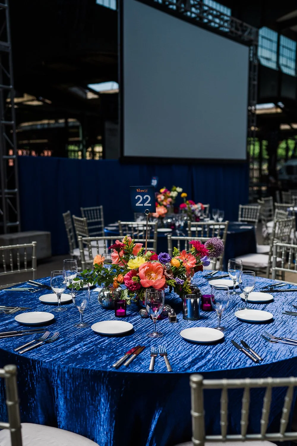 Round banquet table with a blue textured tablecloth, floral centerpiece, white plates, silverware, wine glasses, purple candles, and a blue table number sign, set for an event in a large, industrial-style venue with a blank large screen.