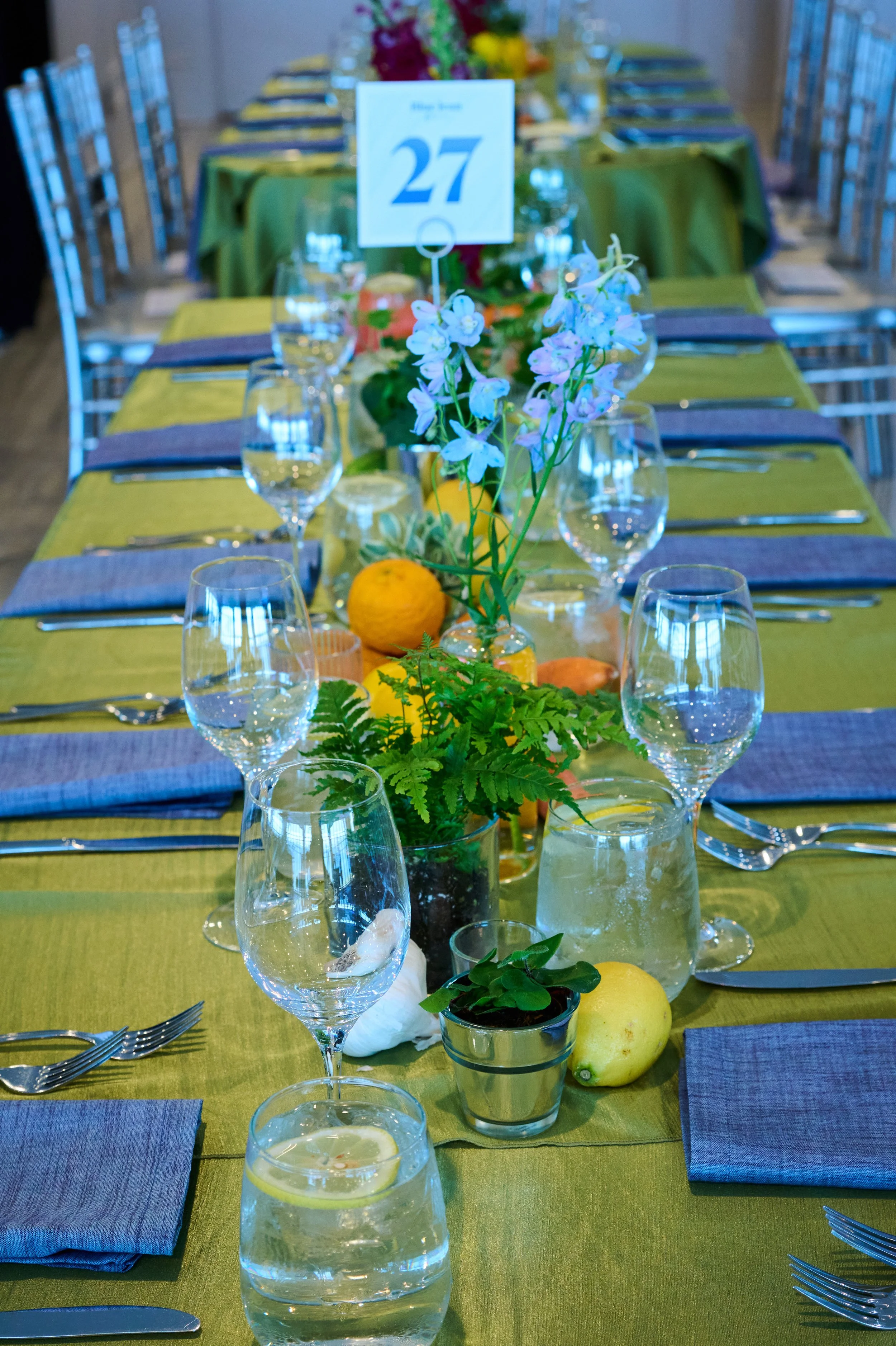 A long dining table set for a formal event with a green tablecloth, gray napkins, multiple wine glasses, and water glasses. The table has floral centerpieces with white and purple flowers, along with lemons and oranges as decoration.