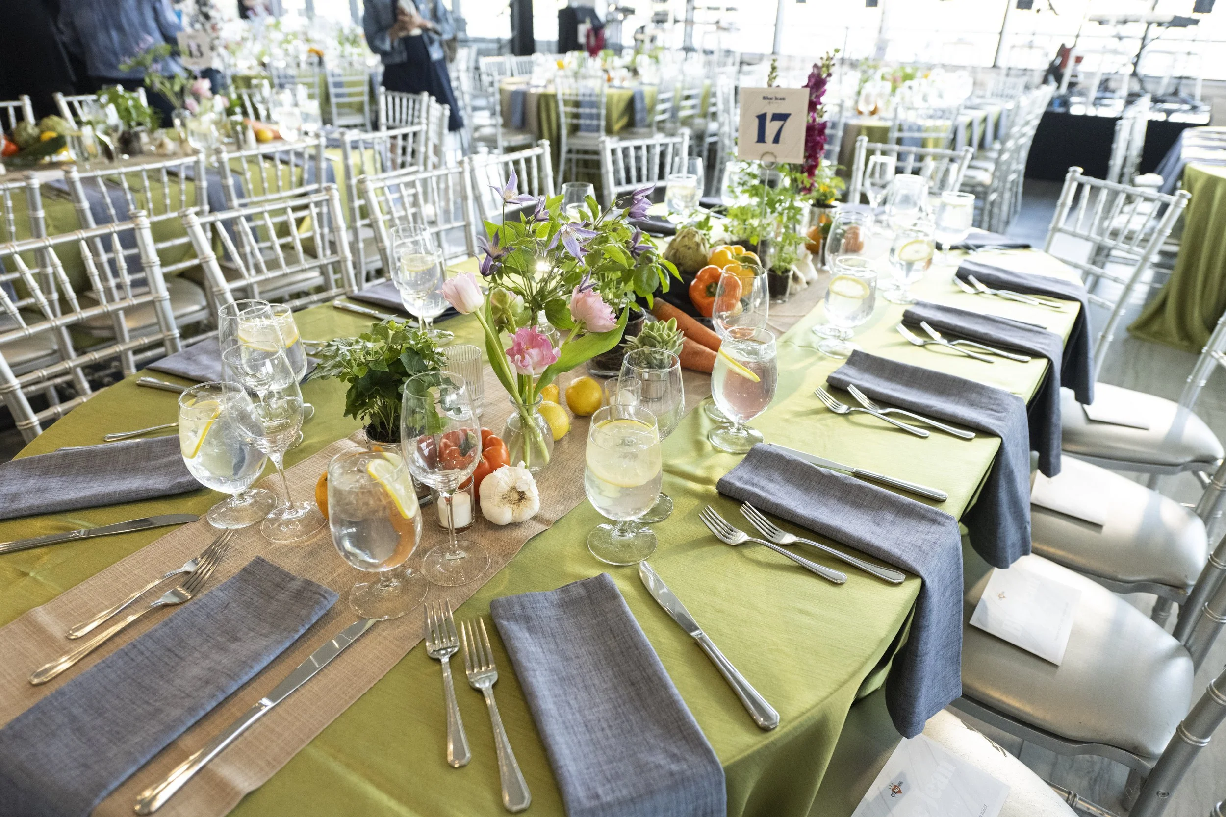 A long banquet table set for a formal event with green tablecloths, gray napkins, and silverware. The table has floral centerpieces with pink and purple flowers and decorative fruits and vegetables. Glasses of water with lemon slices are placed at ea