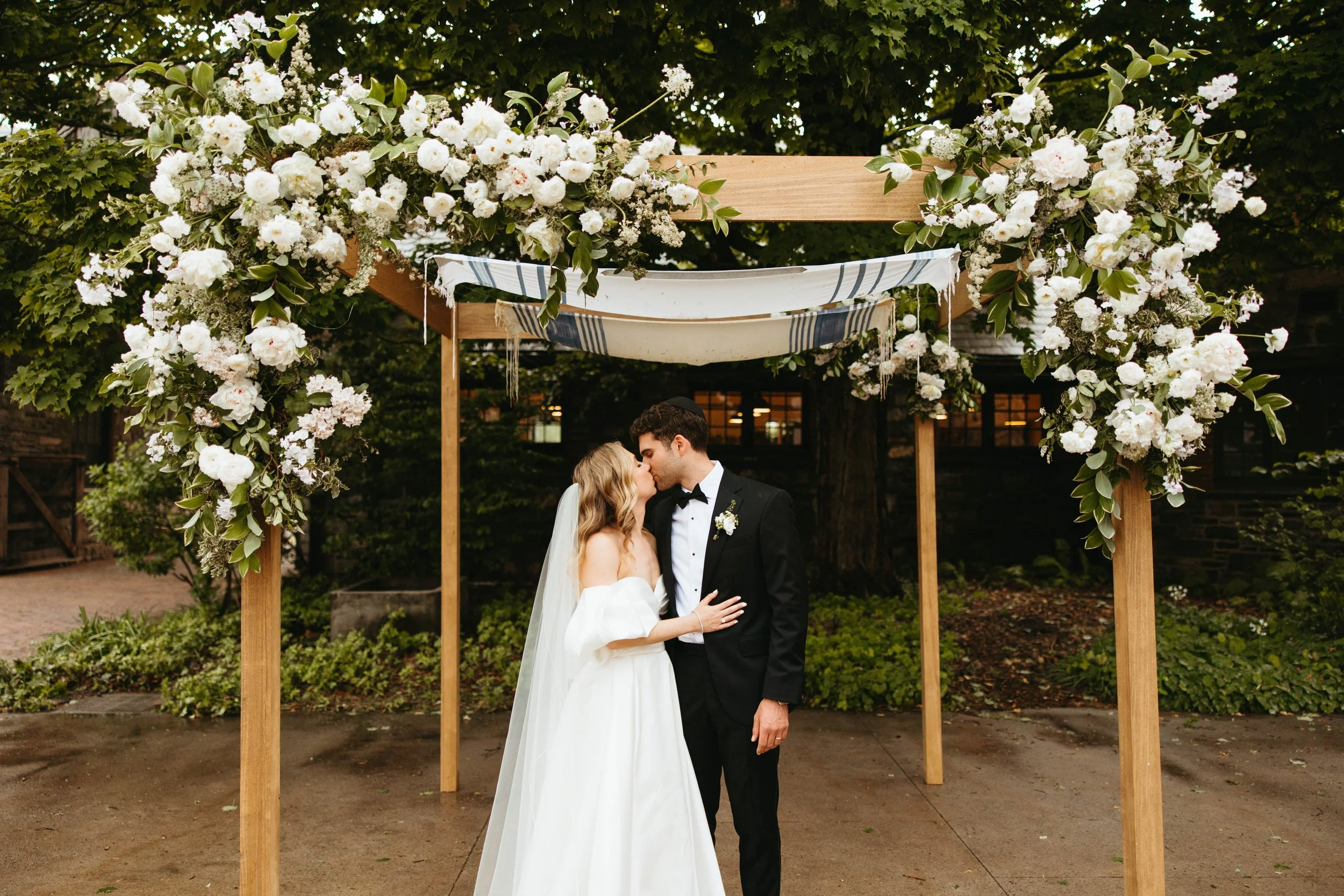 A bride and groom kissing under a floral wedding arch outdoors.
