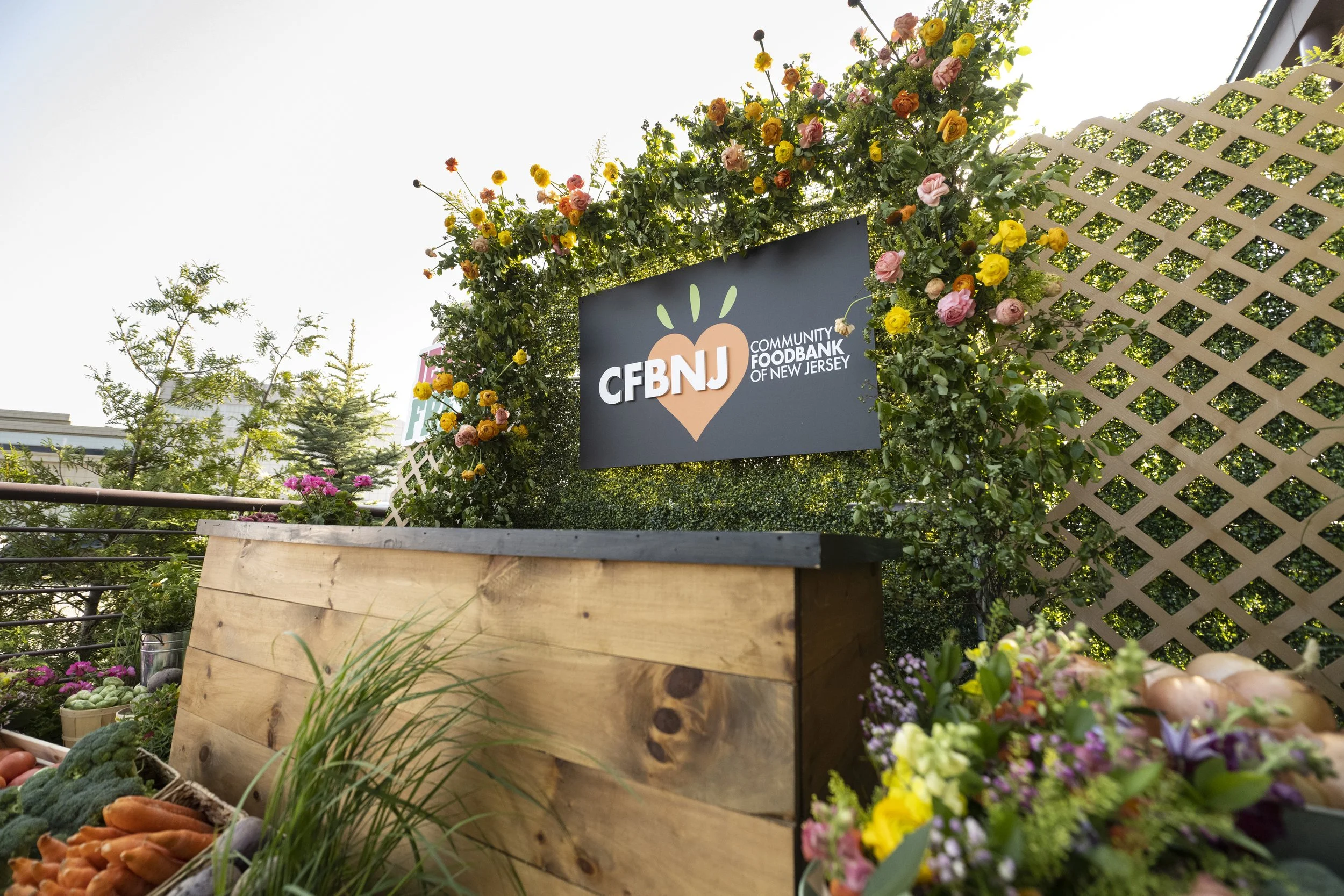 A flower stand or booth decorated with a floral arch, displaying a sign that reads 'CFBNJ' and 'Community Foodbank of New Jersey'. The stand has fresh vegetables like carrots and squashes, with lush green plants and trees in the background.