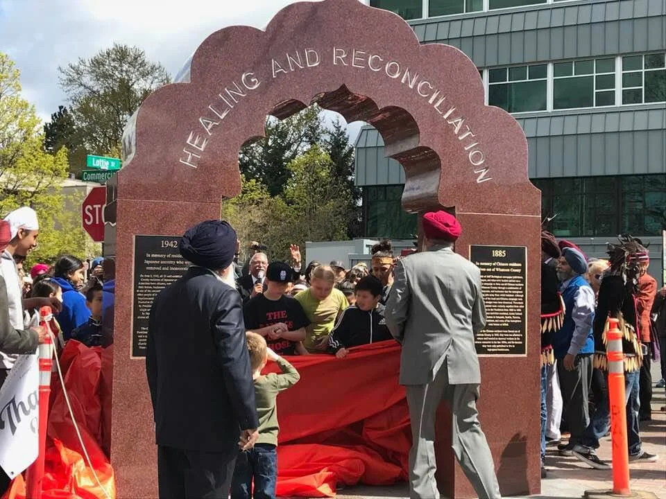 A gathering of people of all ages stand around The Arch of Healing and Reconciliation, a stone archway made of solid red granite