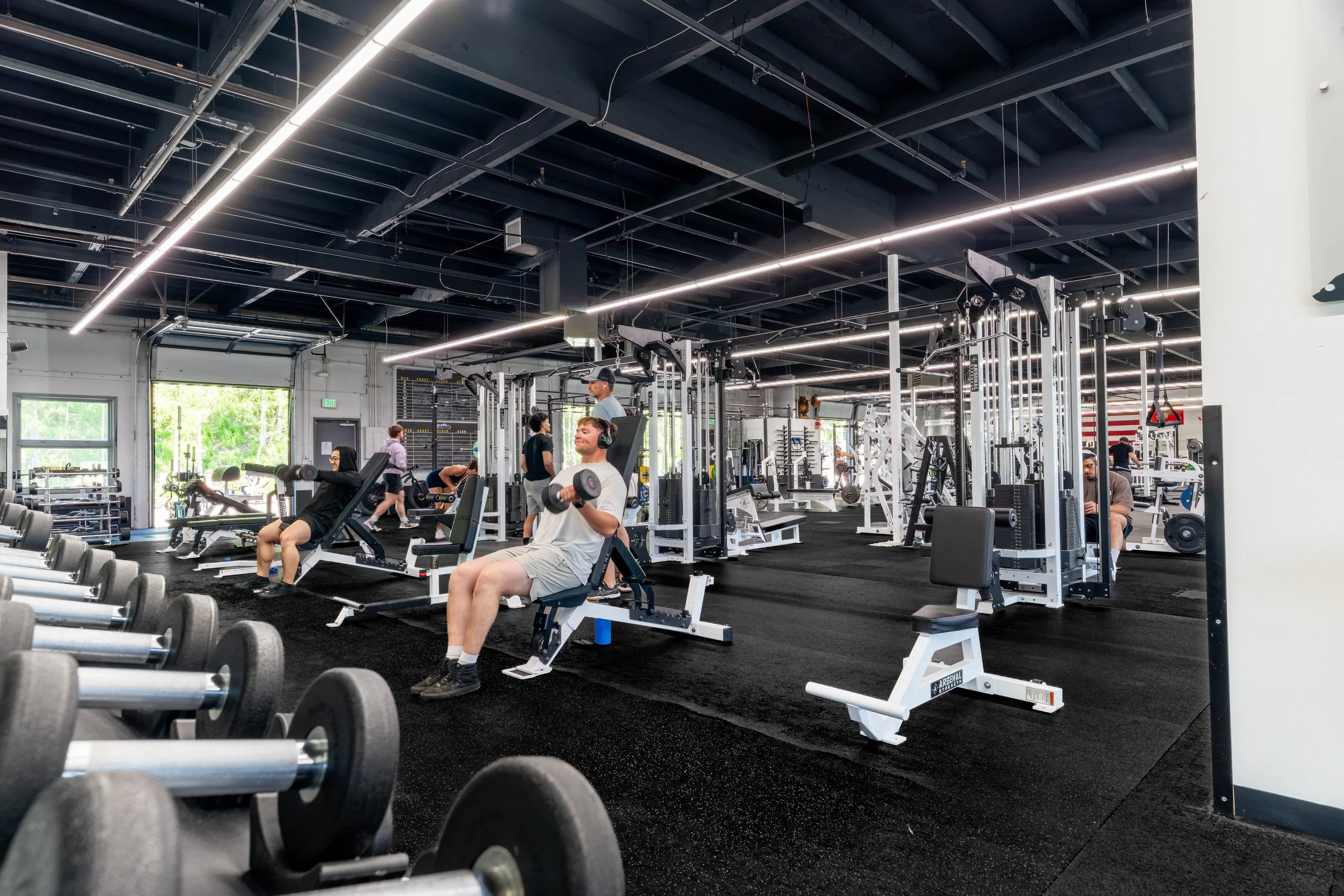 Interior of a modern gym with people working out, including individuals using weight machines and lifting dumbbells.
