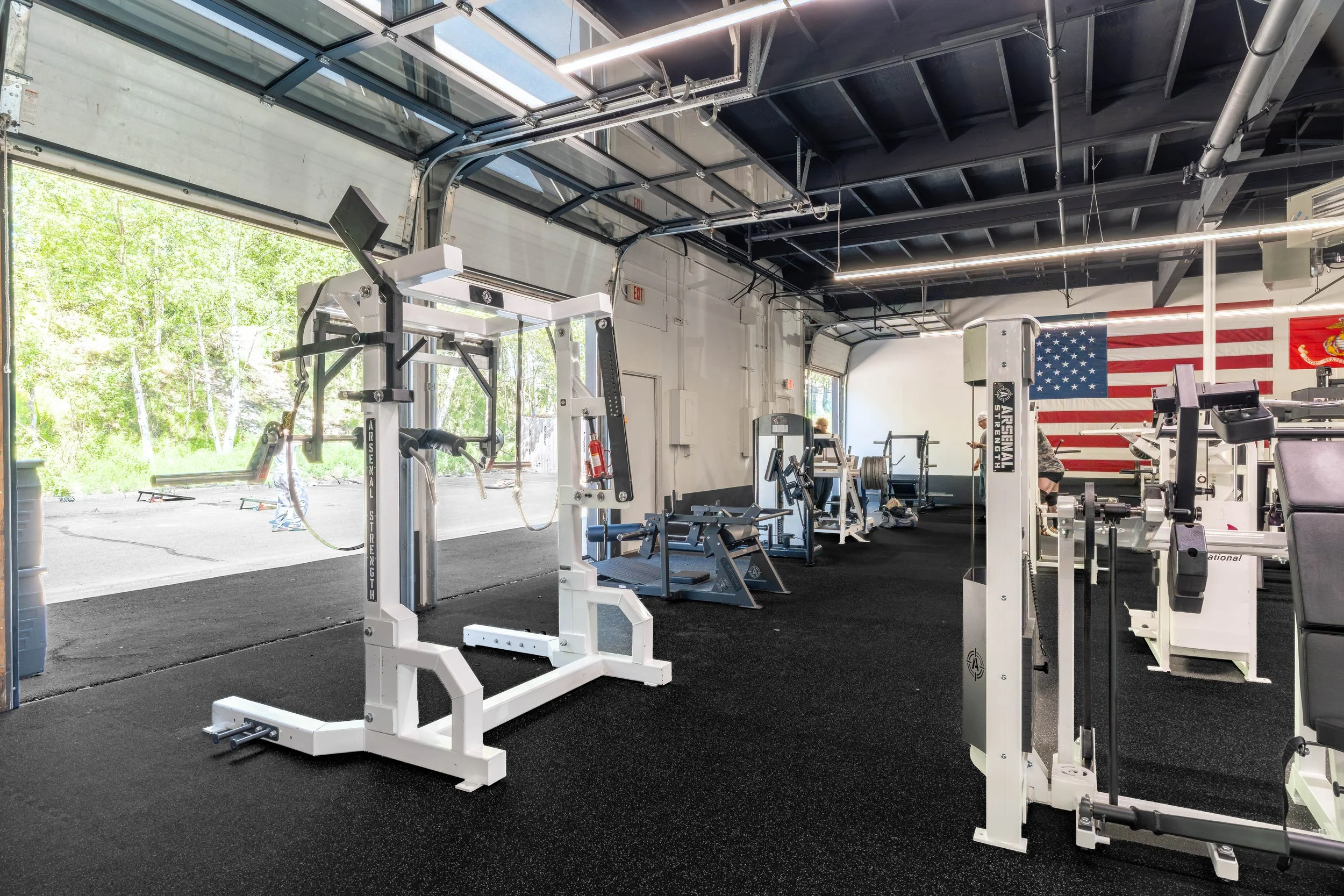 Inside a gym with fitness equipment, large American flag on wall, open garage-style door showing green trees outside, black rubber flooring, and ceiling with exposed beams and lighting.