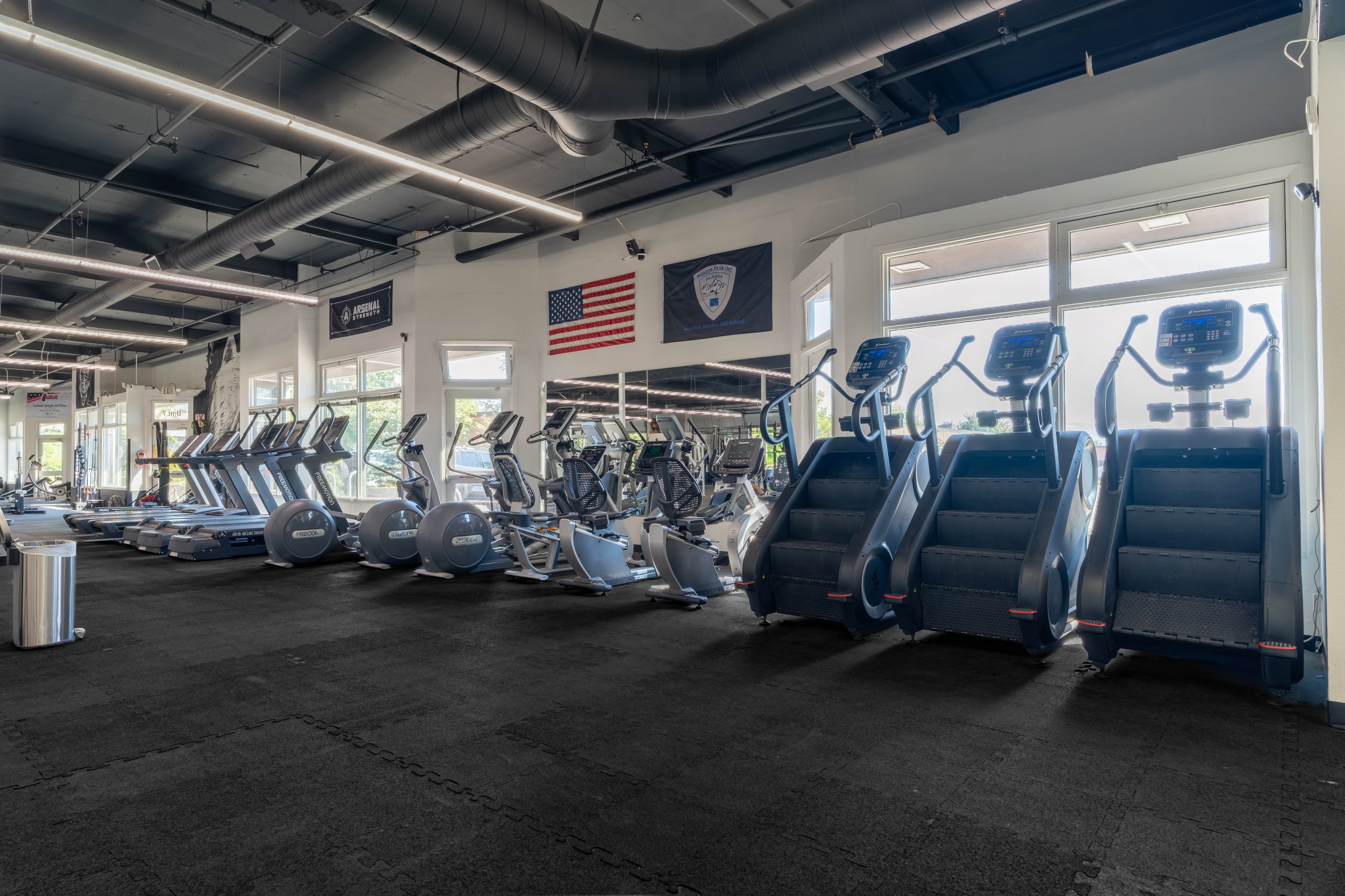Interior of a gym with various exercise equipment including treadmills, elliptical machines, and stair climbers, with large windows and flags hanging on the wall.