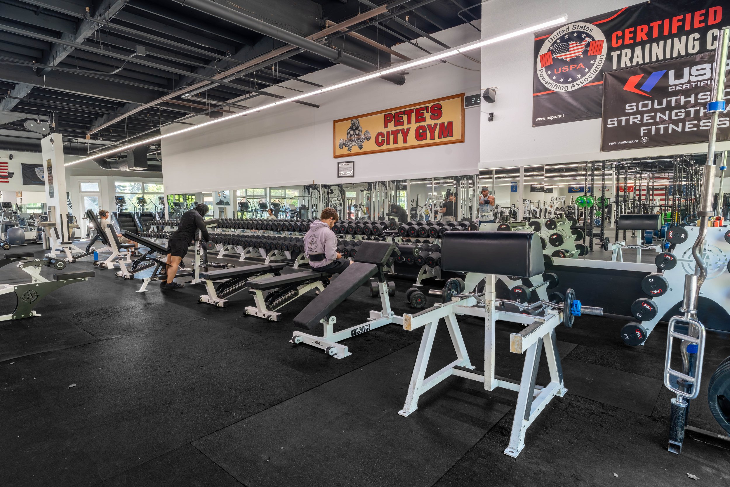 Interior of a gym with various exercise equipment, people working out, and signage including 'Pete's City Gym' and banners for USPA certifications.