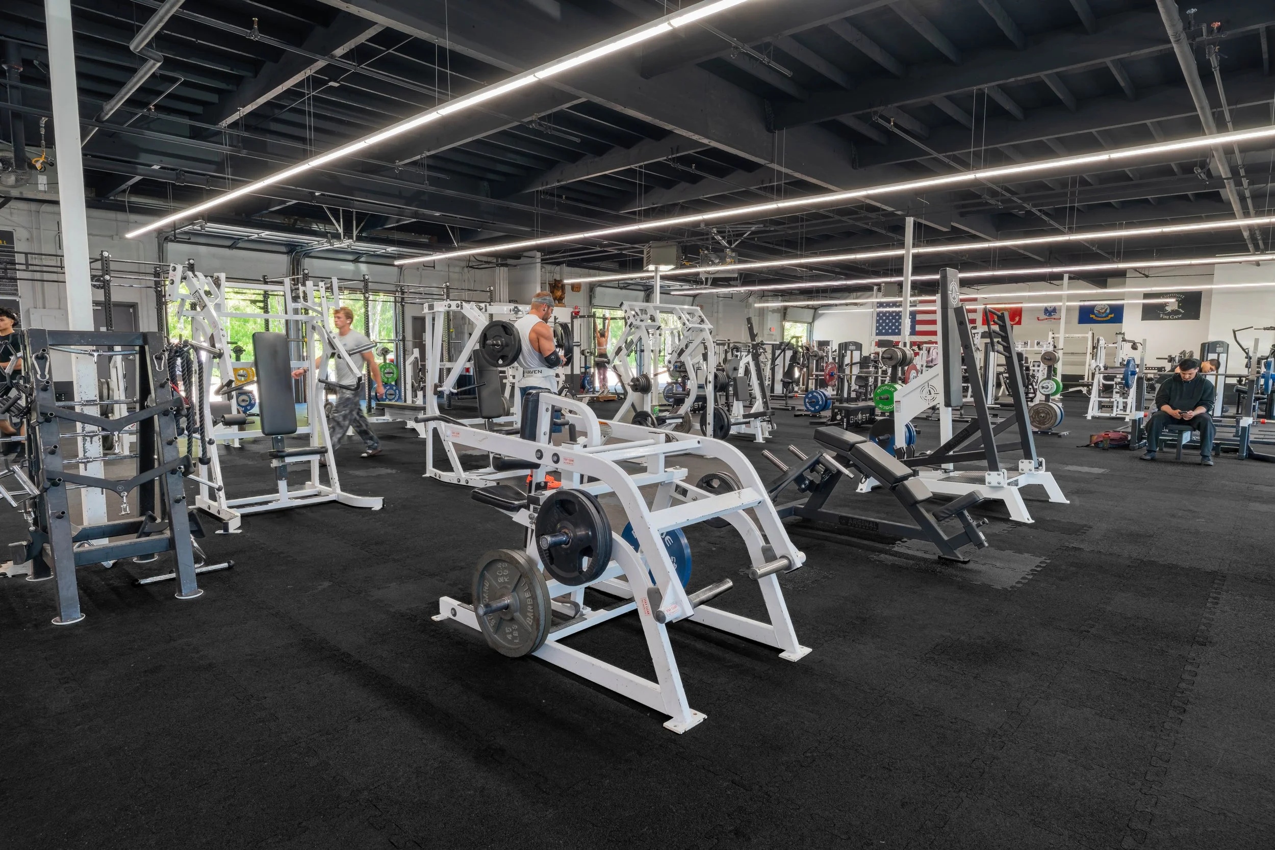 A gym with various workout equipment, including weight machines and free weights. Several people are working out or sitting on benches, with American flags and gym banners in the background.
