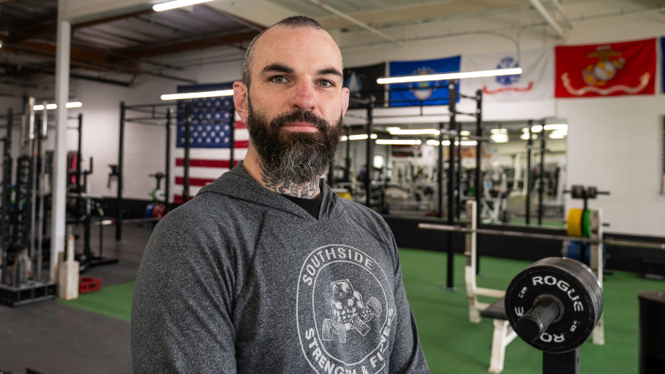A bearded man with short hair standing in a gym, wearing a gray hooded sweatshirt, with weightlifting equipment and American, marine, and navy flags in the background.