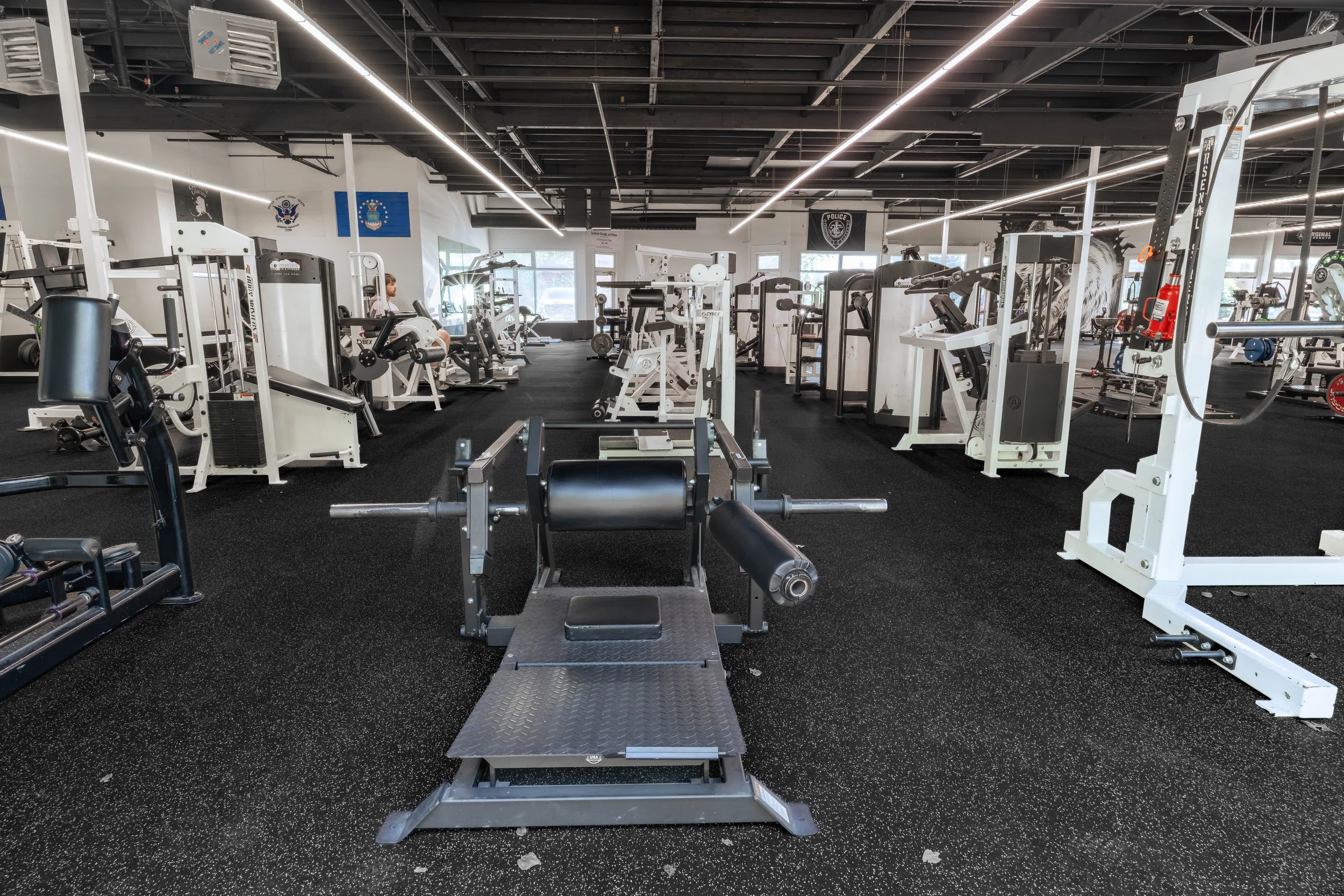 A spacious modern gym with various exercise equipment, including weight machines, benches, and cardio machines, with large windows and flags on the walls.