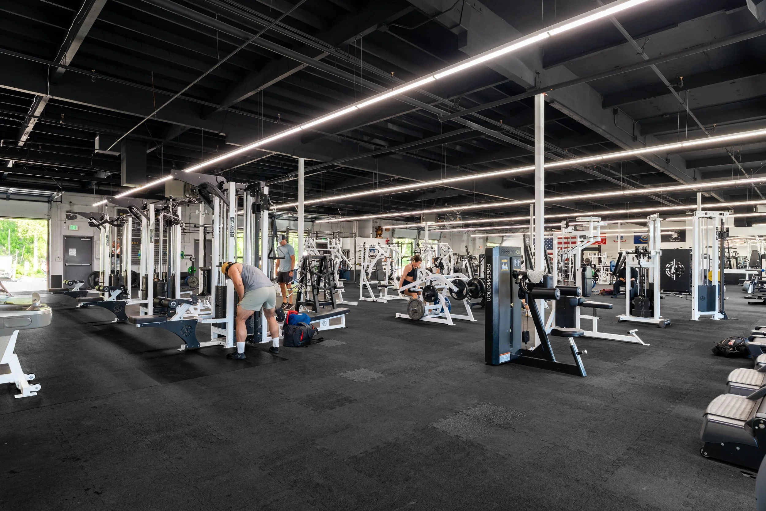 People working out with gym equipment inside a spacious fitness center with black walls and ceiling, illuminated by long ceiling lights.