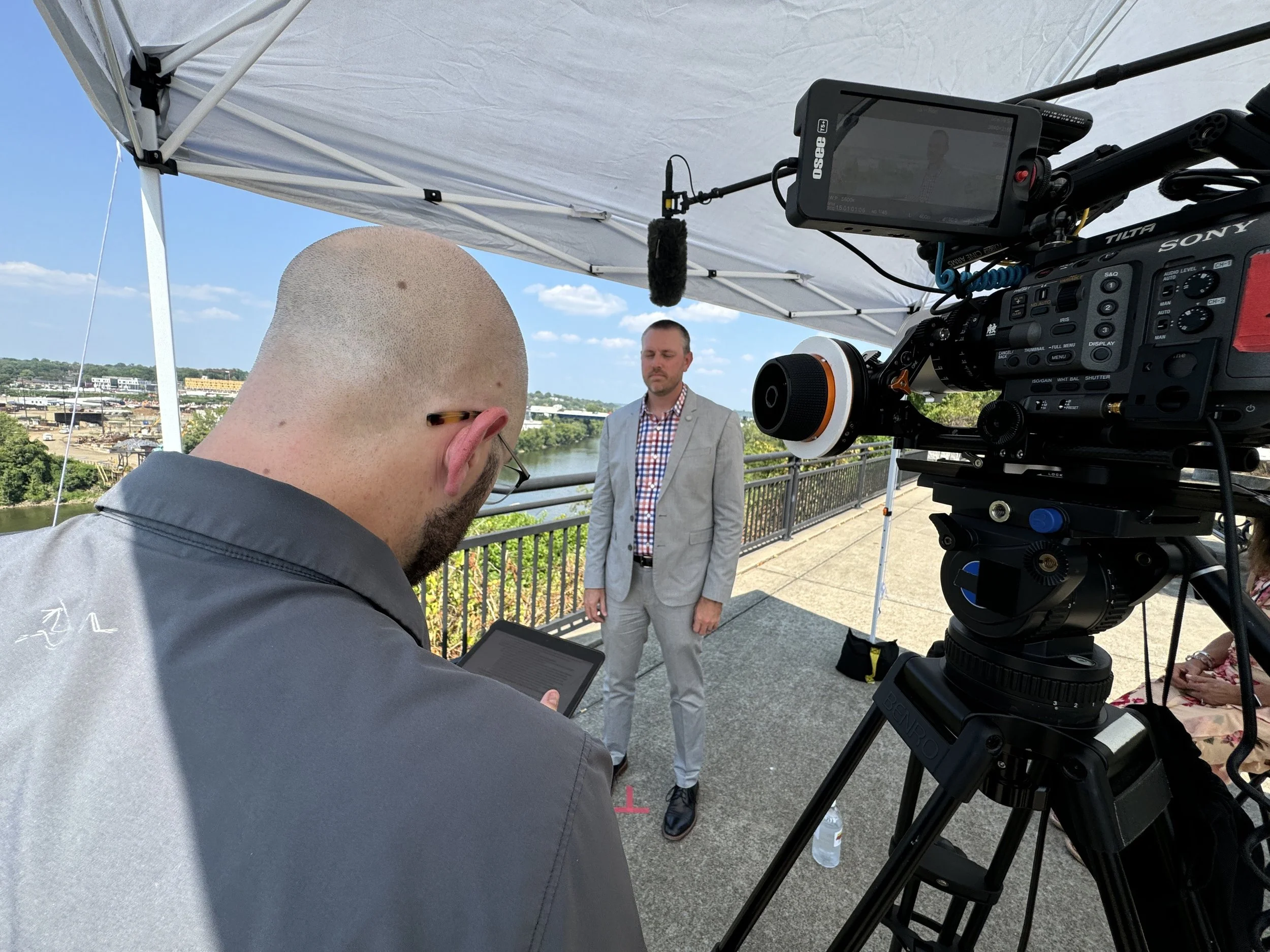 Director Nick prepares for an outdoor interview with Kent Archer on a riverfront overlook, framed by a Sony cinema camera and shaded by a tent during a Stantec flood resilience film shoot.