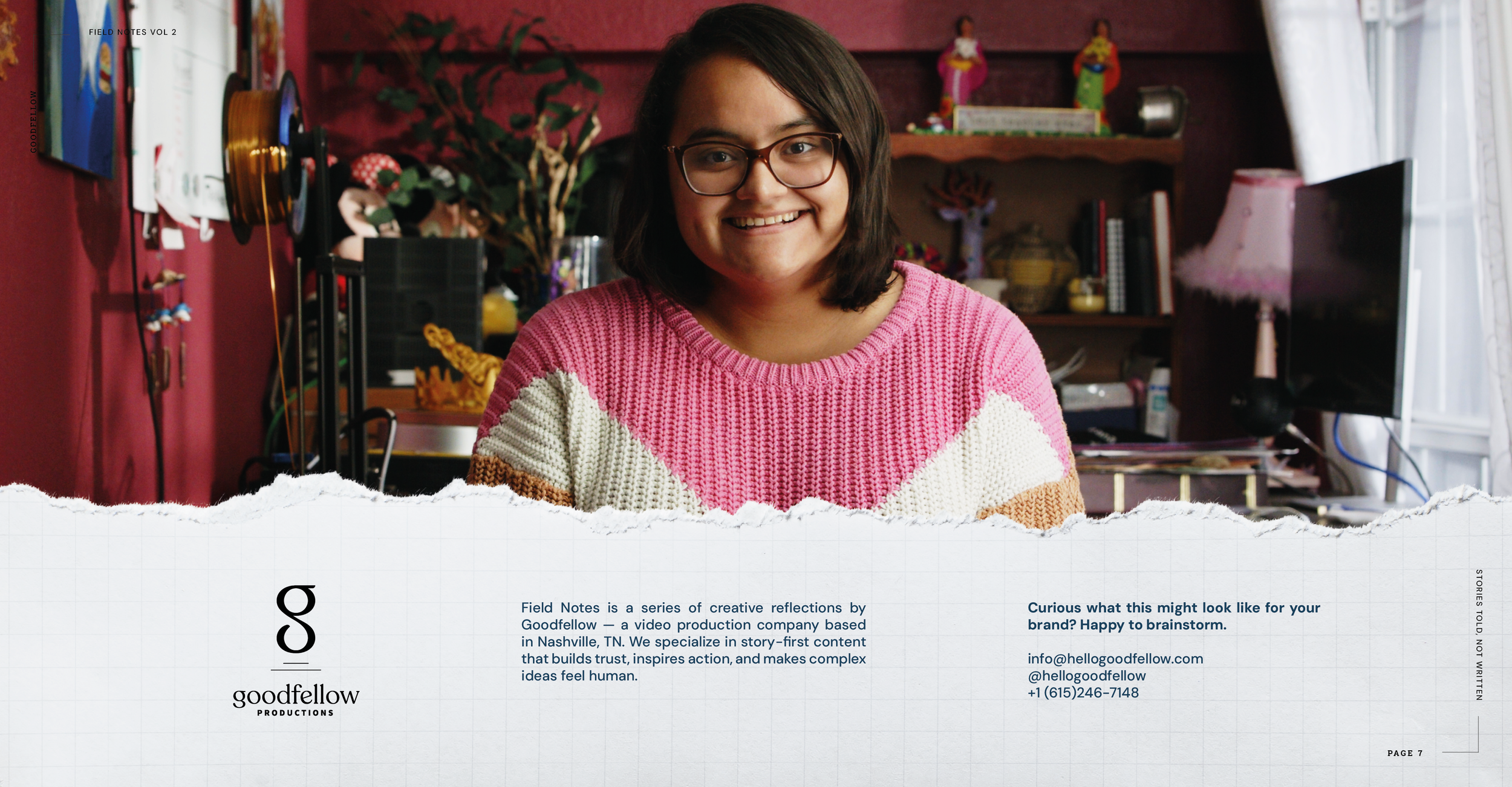 A young woman with dark hair, glasses, and a pink and cream sweater, smiling in a colorful, cluttered room with shelves, a lamp, and a computer