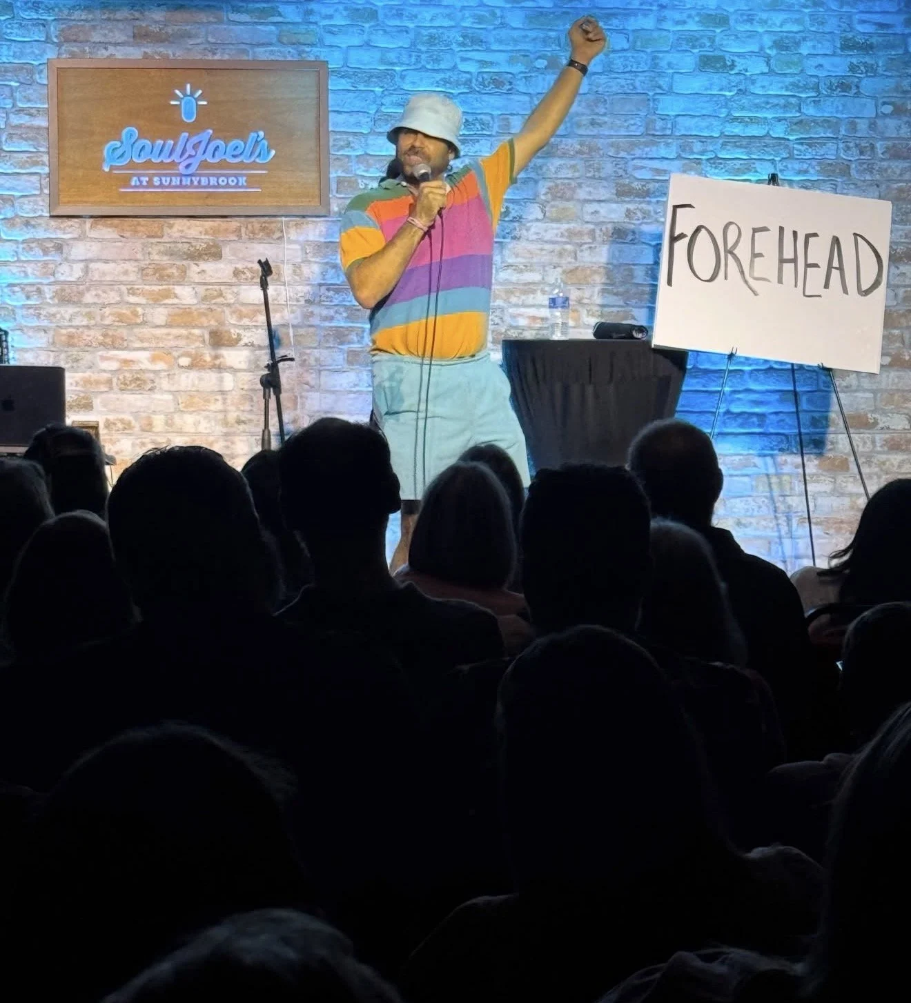 A comedian on stage at the Ludlow's comedy club, wearing a colorful striped shirt, beige bucket hat, and holding a microphone, raising one hand. A large sign next to him says 'Forehead'. The audience is seated in front of him.