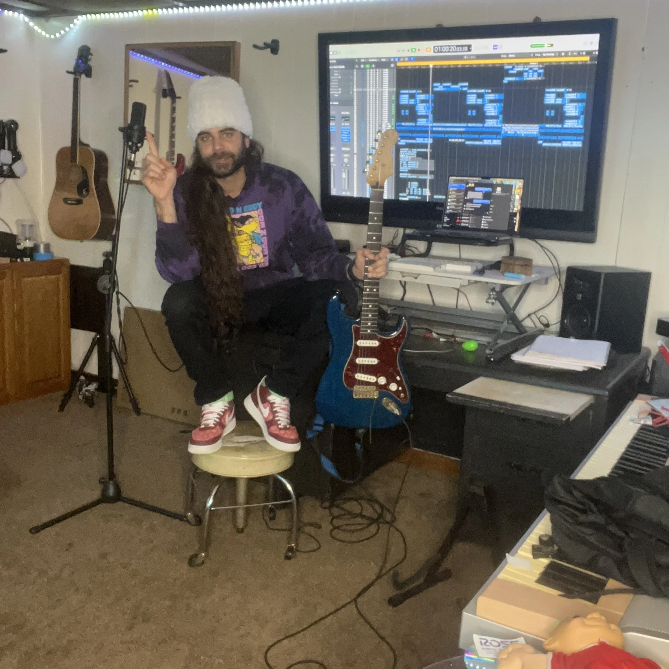 A man with long hair and a beard wearing a fluffy white hat and a graphic purple shirt, holding a guitar, standing in a music studio with guitars hanging on the wall, a large computer monitor displaying a digital audio workstation, and musical instruments on a desk.