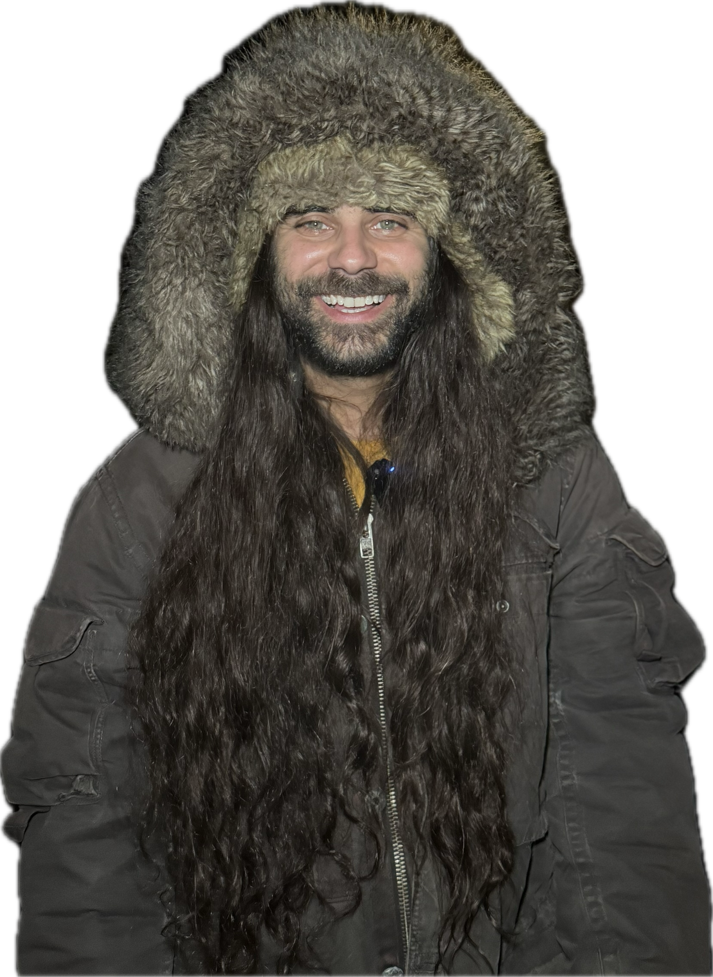 A smiling man with long dark hair and a beard wearing a large, fluffy fur-lined hooded winter coat against a black background.