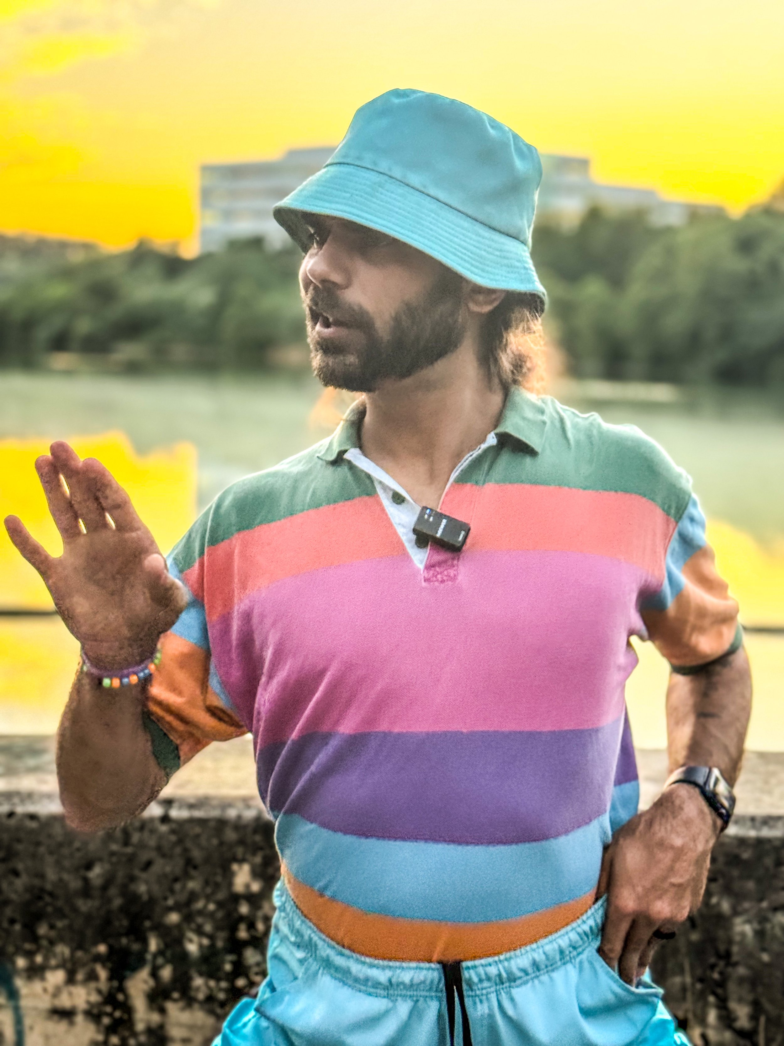 A man wearing a turquoise bucket hat, a colorful striped polo shirt, and turquoise shorts, standing outdoors near a body of water during sunset, gesturing with his right hand.