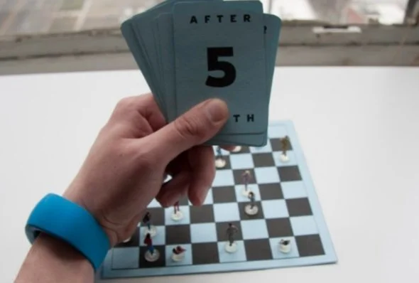 Person holding a card with the number 5 above a chessboard with chess pieces set up for a game.